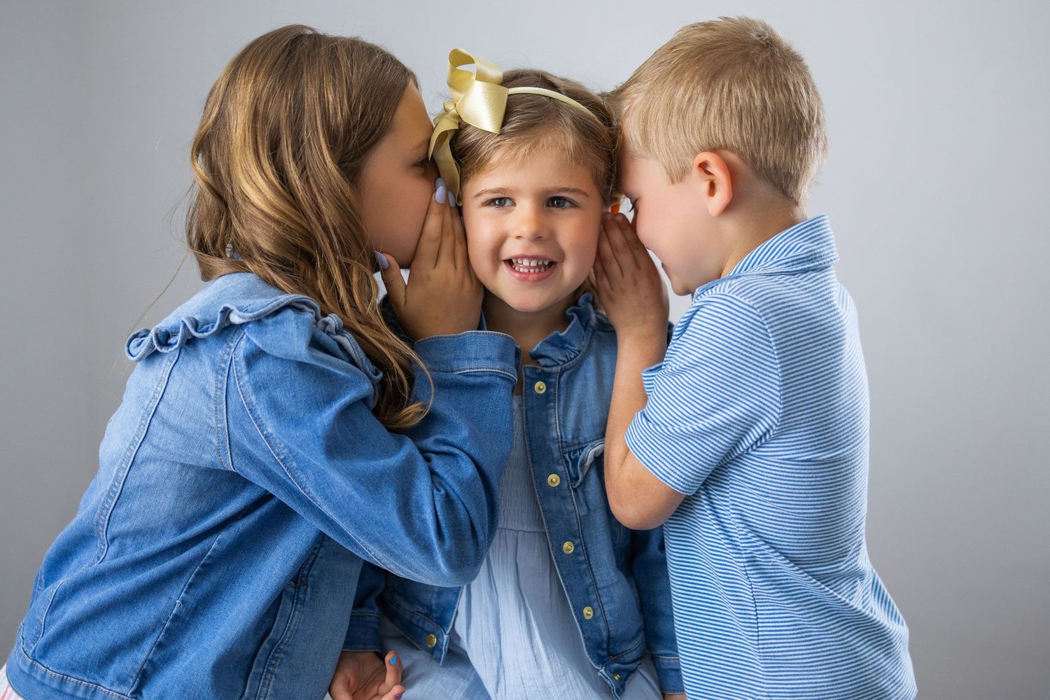 Three children in denim outfits share secrets and giggles against a gray background.