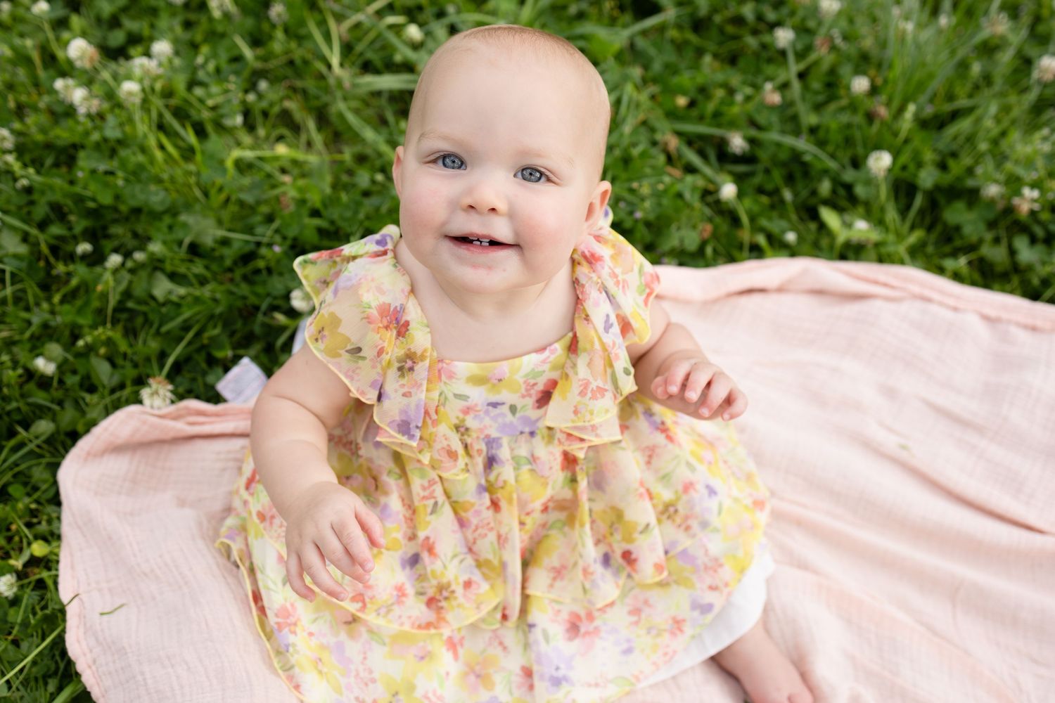 Baby in floral yellow dress sitting on pink blanket outdoors.