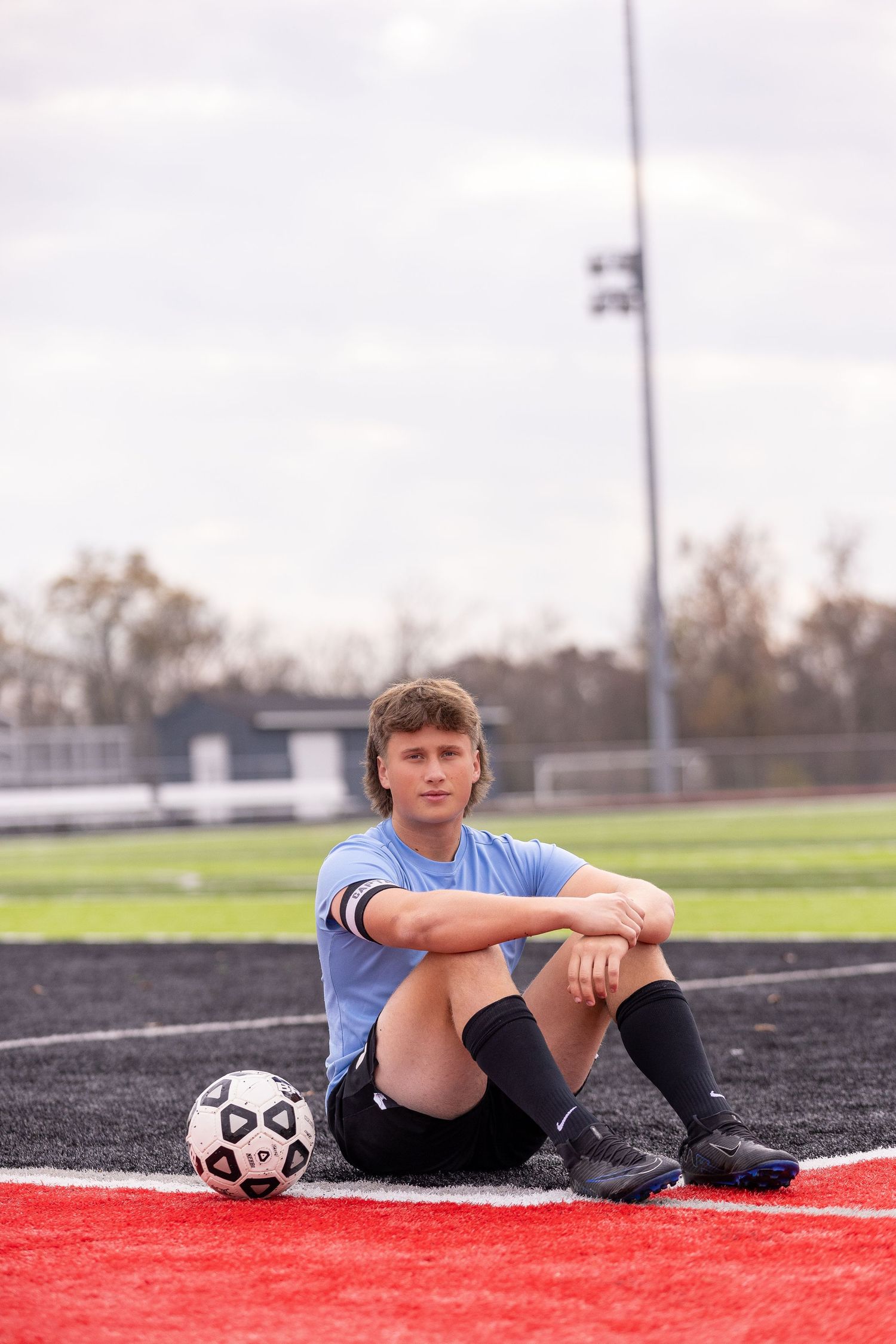 Soccer player sitting on track field with ball wearing light blue jersey and knee socks.