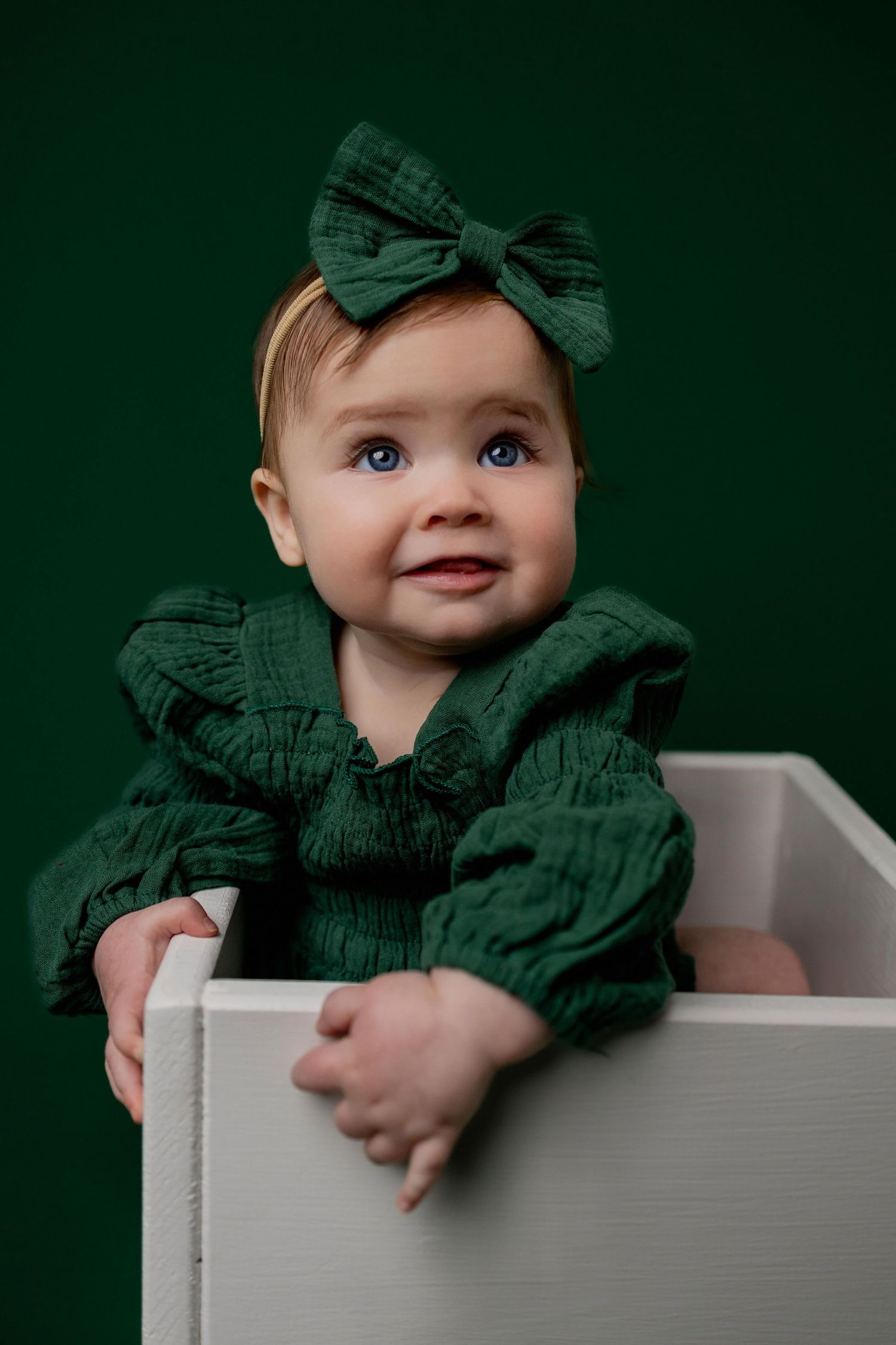 Adorable baby wearing forest green outfit with bow sits in white box against dark green backdrop.