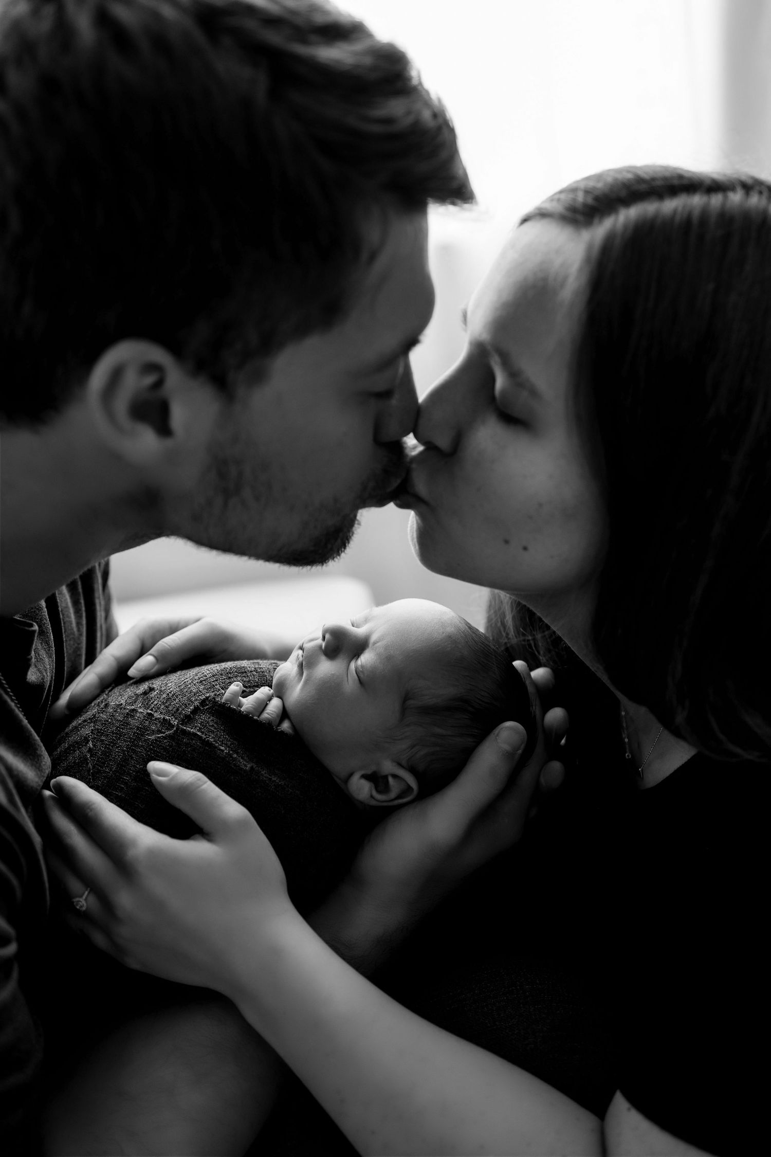 Black and white portrait of parents sharing tender moment with sleeping newborn between them.