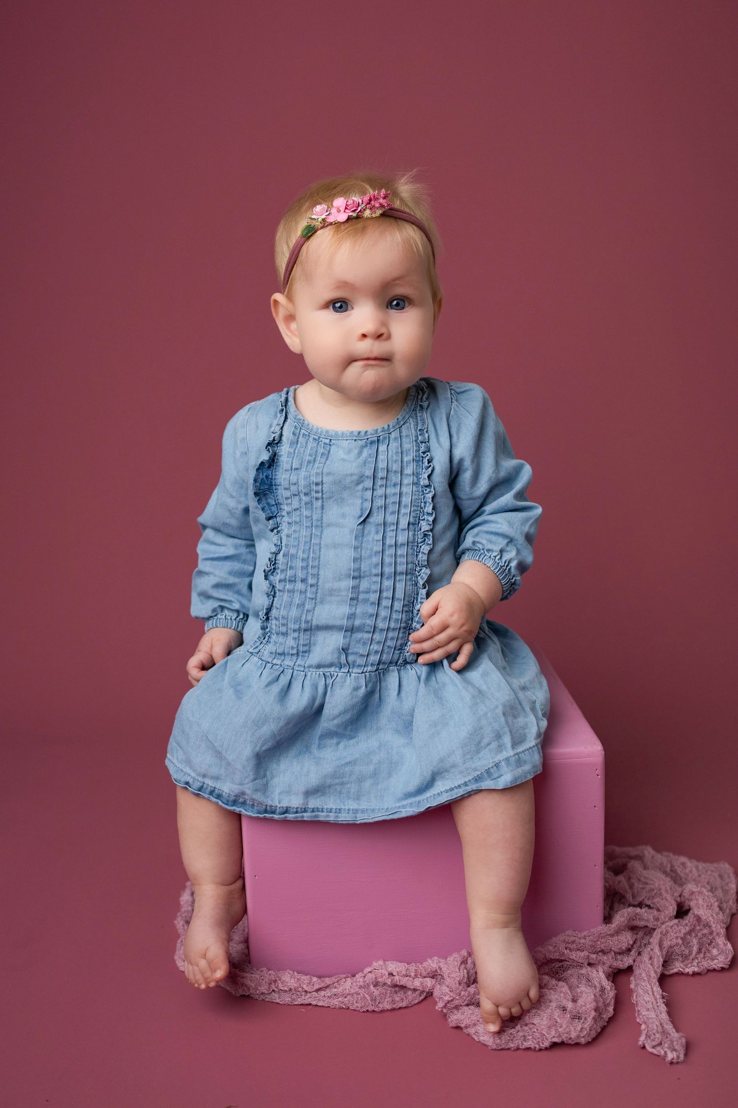 Baby wearing blue denim dress and floral headband sits on pink stool against burgundy background.