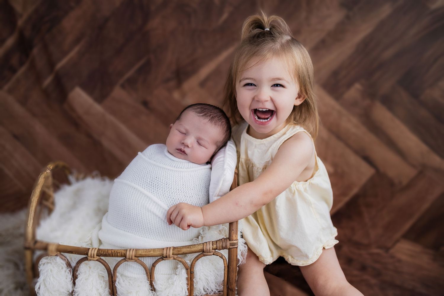 A joyful toddler in a cream dress leans over a wicker bassinet containing a swaddled sleeping infant.