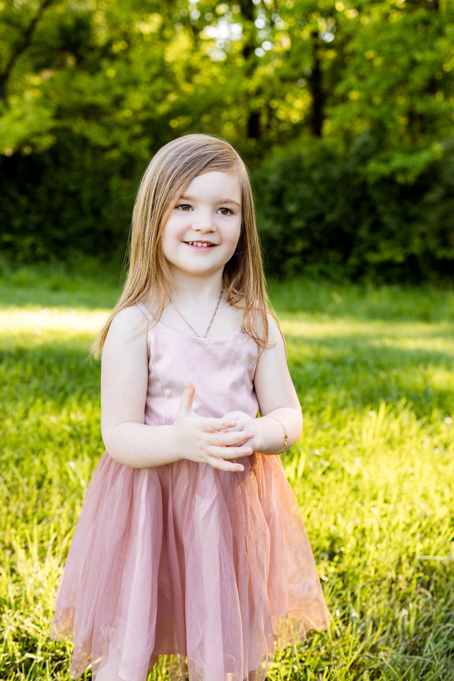 Young child in pink tulle dress smiles sweetly while standing in sunlit meadow.