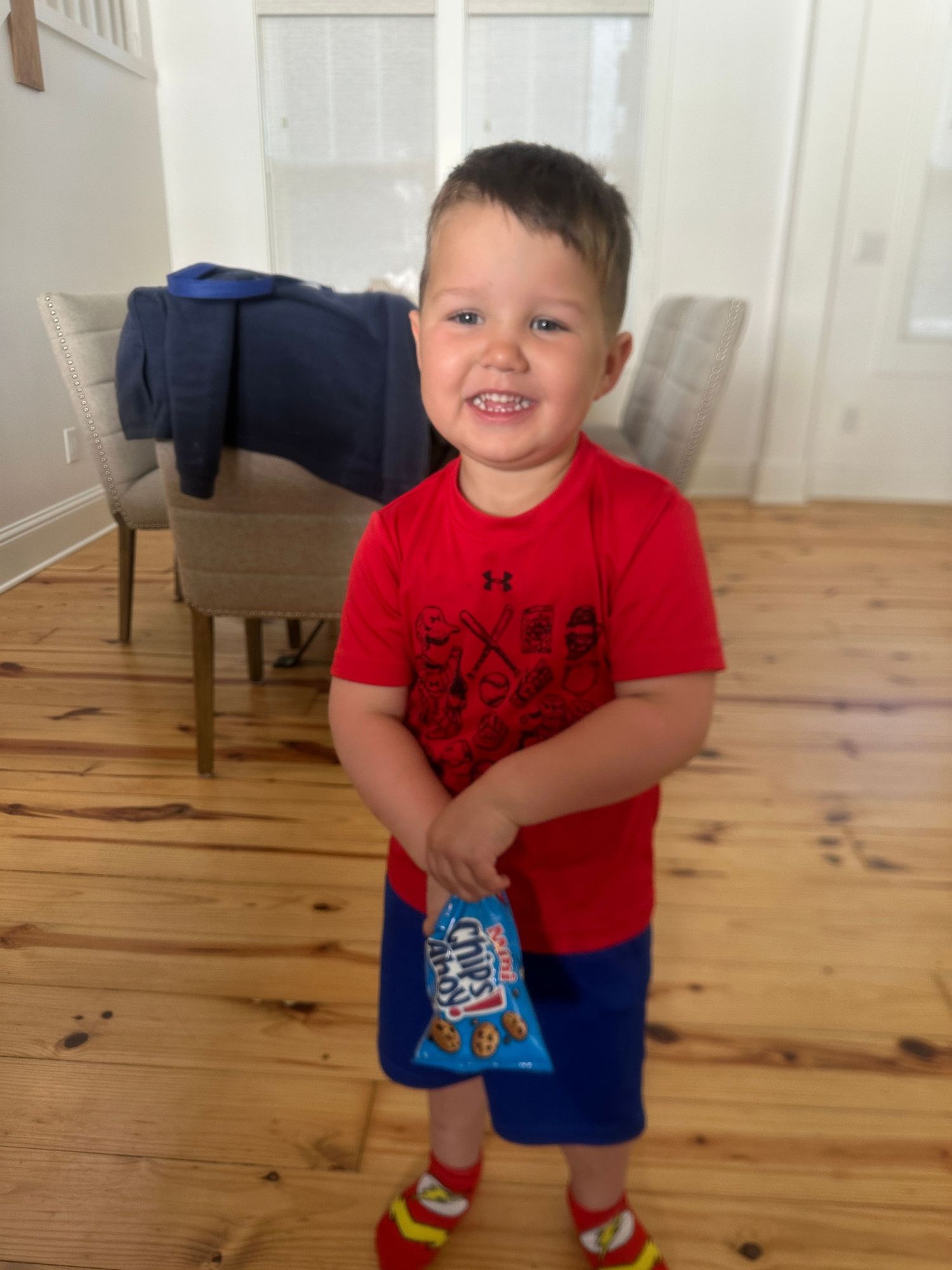Child in red t-shirt and blue shorts smiling in living room with wooden floors.