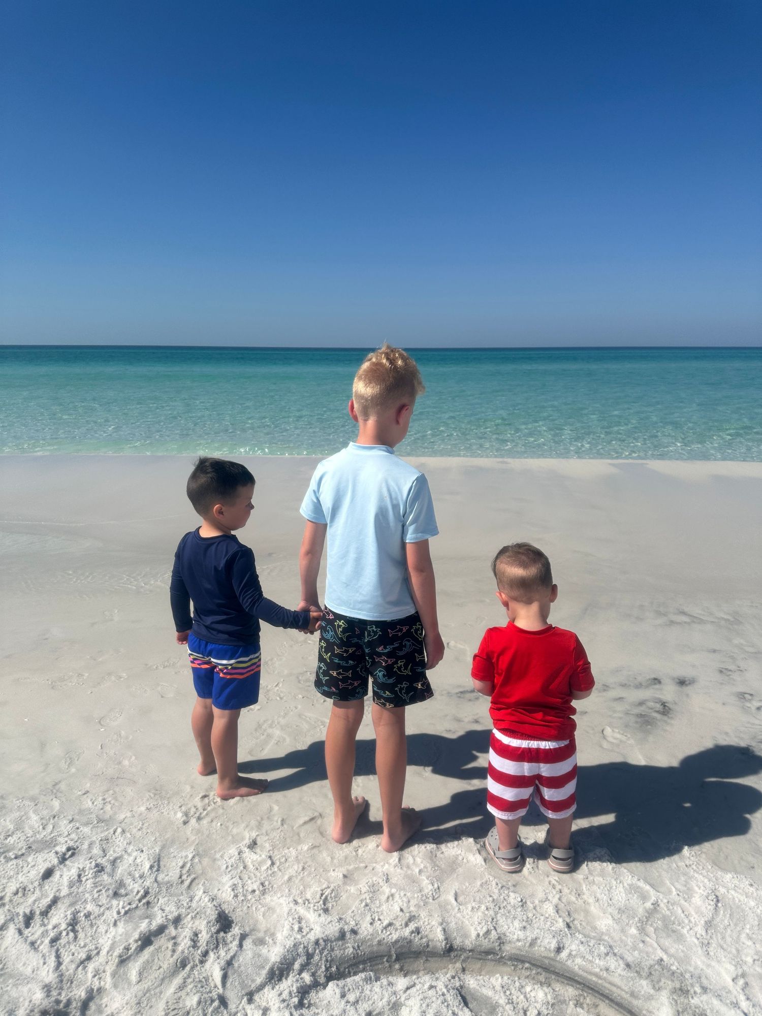 Three children standing at water's edge looking out at turquoise ocean on sunny day.