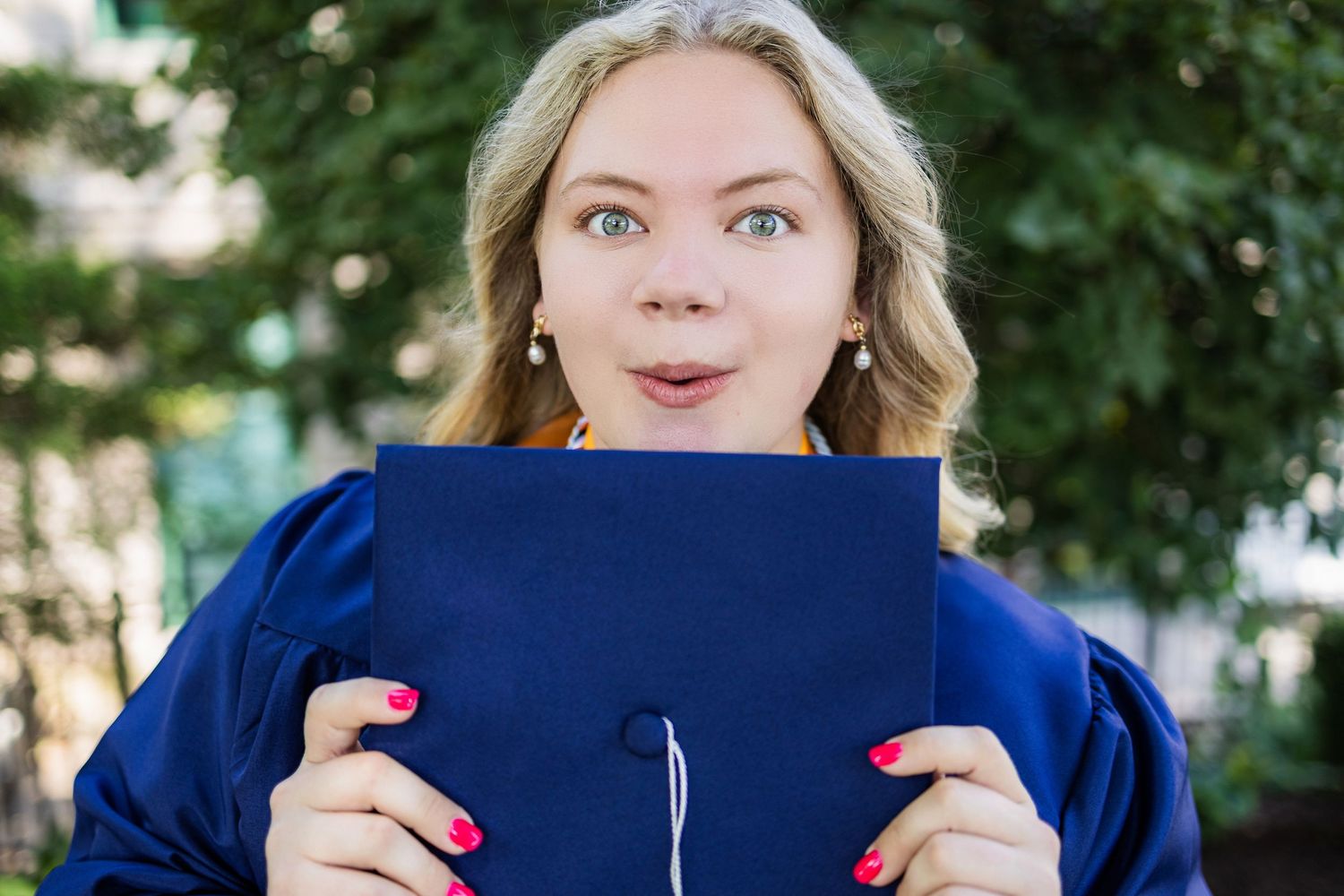 Person with surprised expression holds up a navy blue diploma or graduation folder with bright pink nails outdoors.