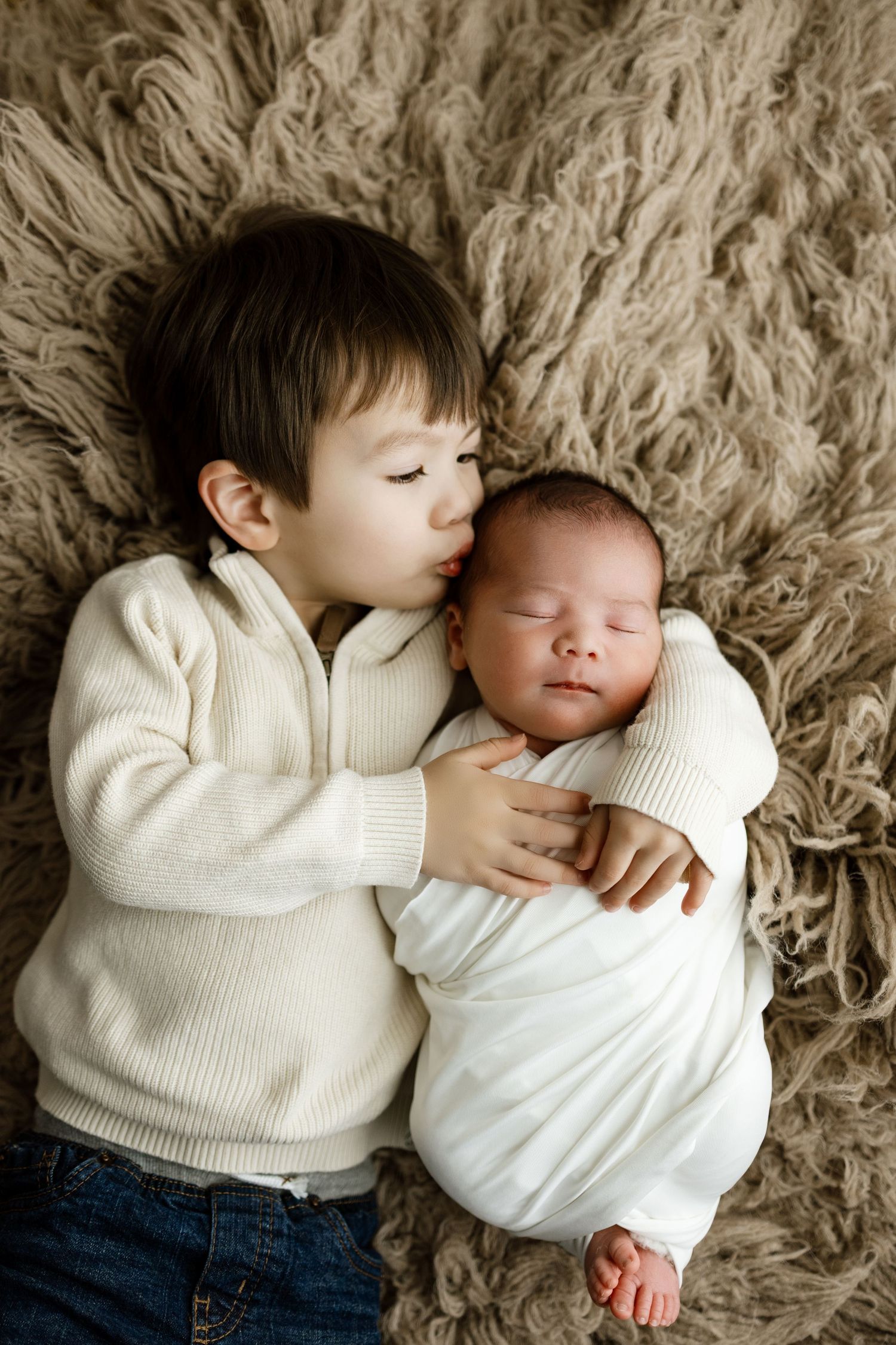 Two siblings in cream sweaters share a tender moment while lying on a fluffy beige carpet.