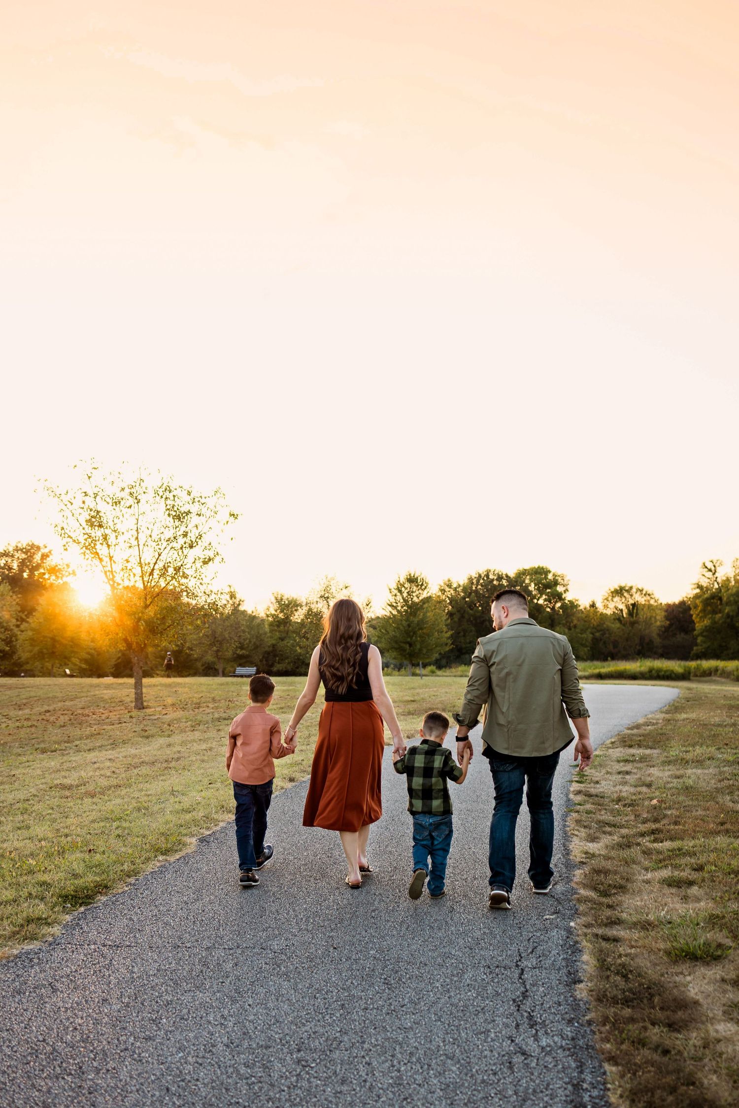 Family walking together on a gravel path at sunset in a rural setting with golden sunlight filtering through trees.