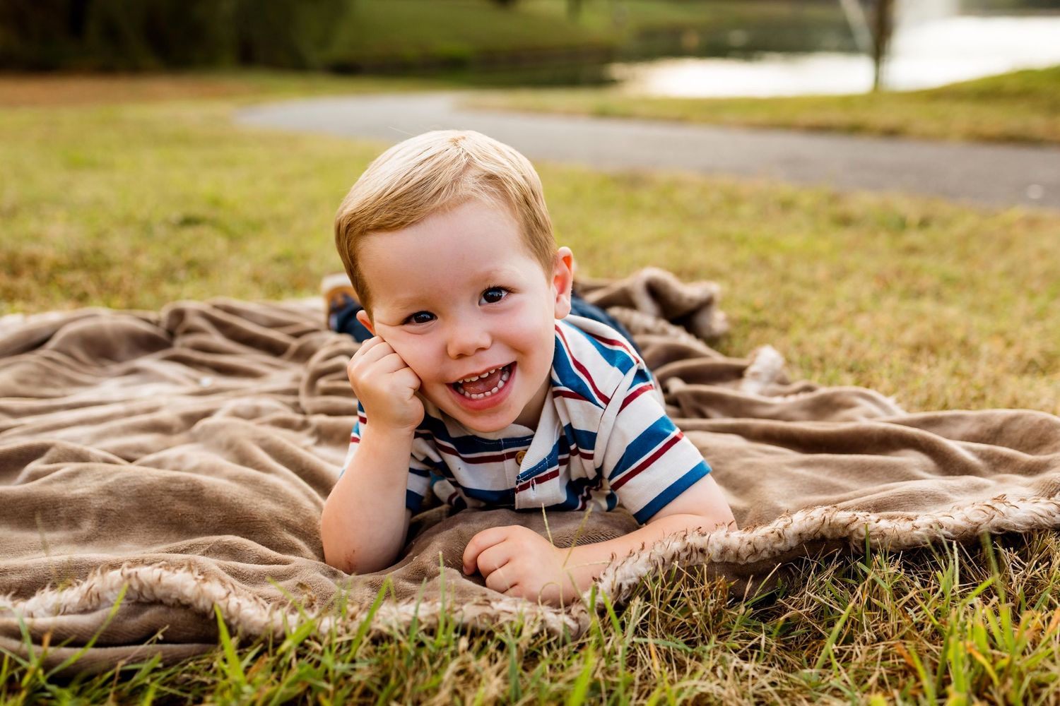 A young child in a striped shirt lies on a brown blanket on grass while smiling at the camera.