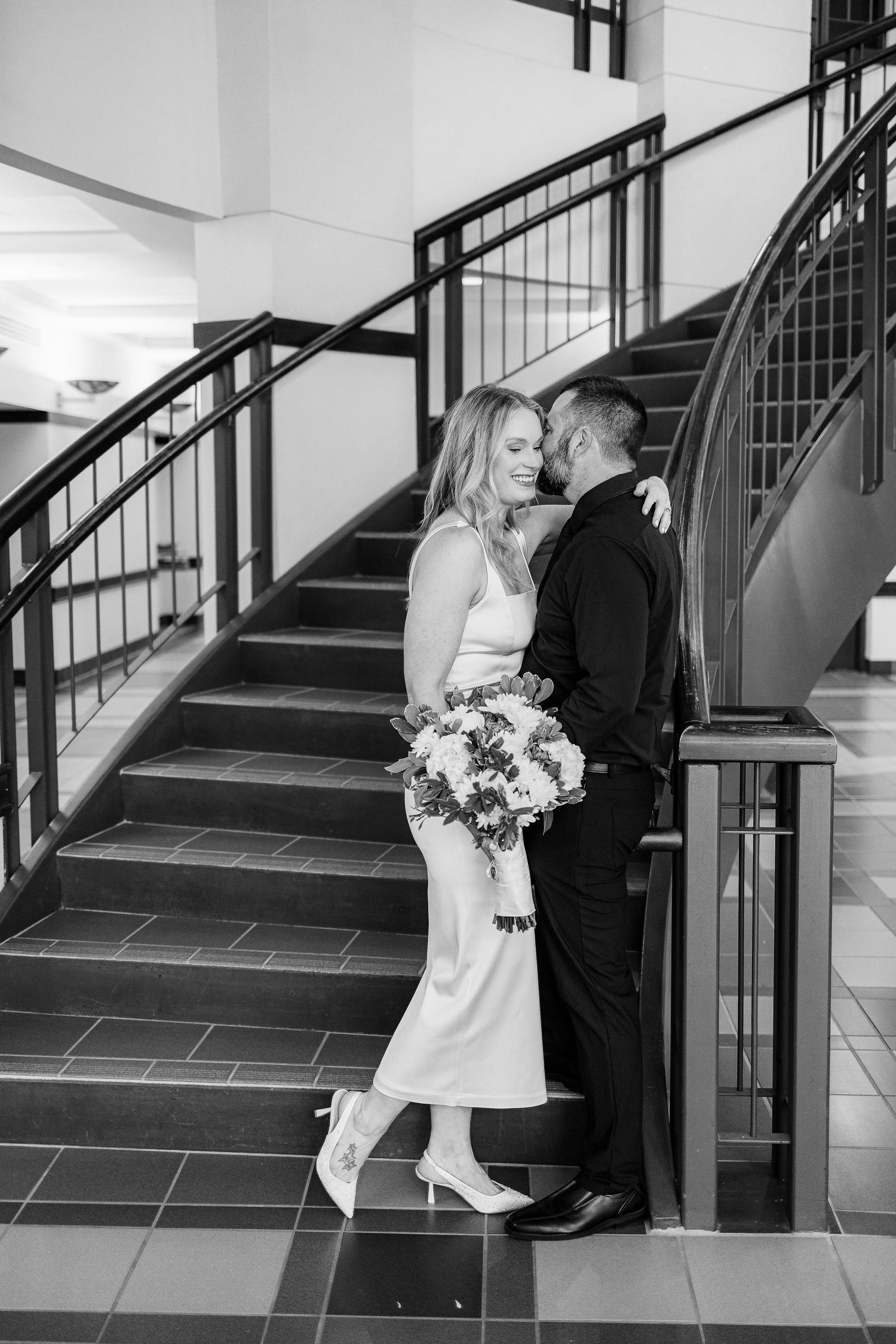 A black and white wedding photo of a couple sharing an intimate moment by a staircase with elegant railings.