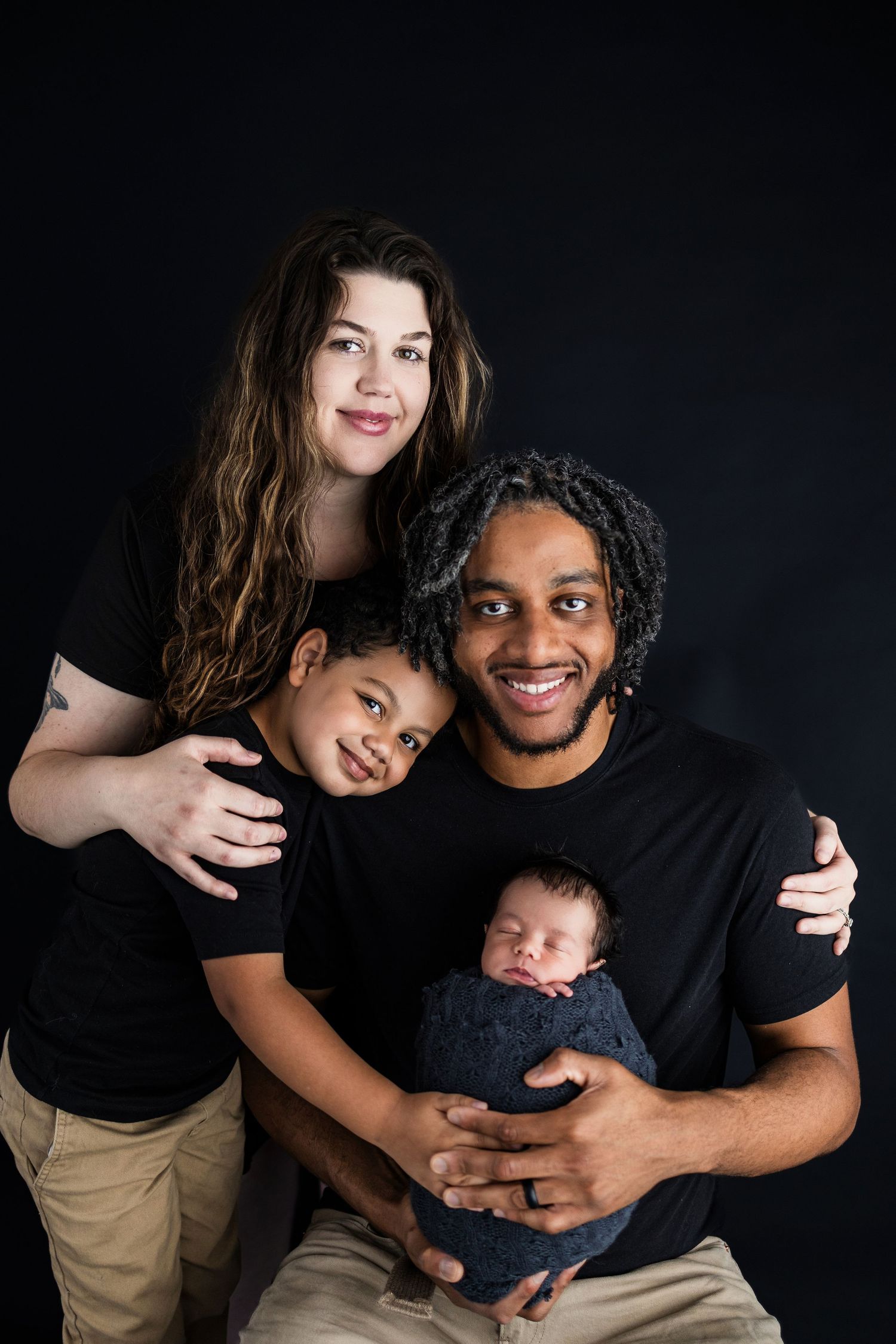 A loving family portrait against a dark background shows parents with their young child and newborn baby.