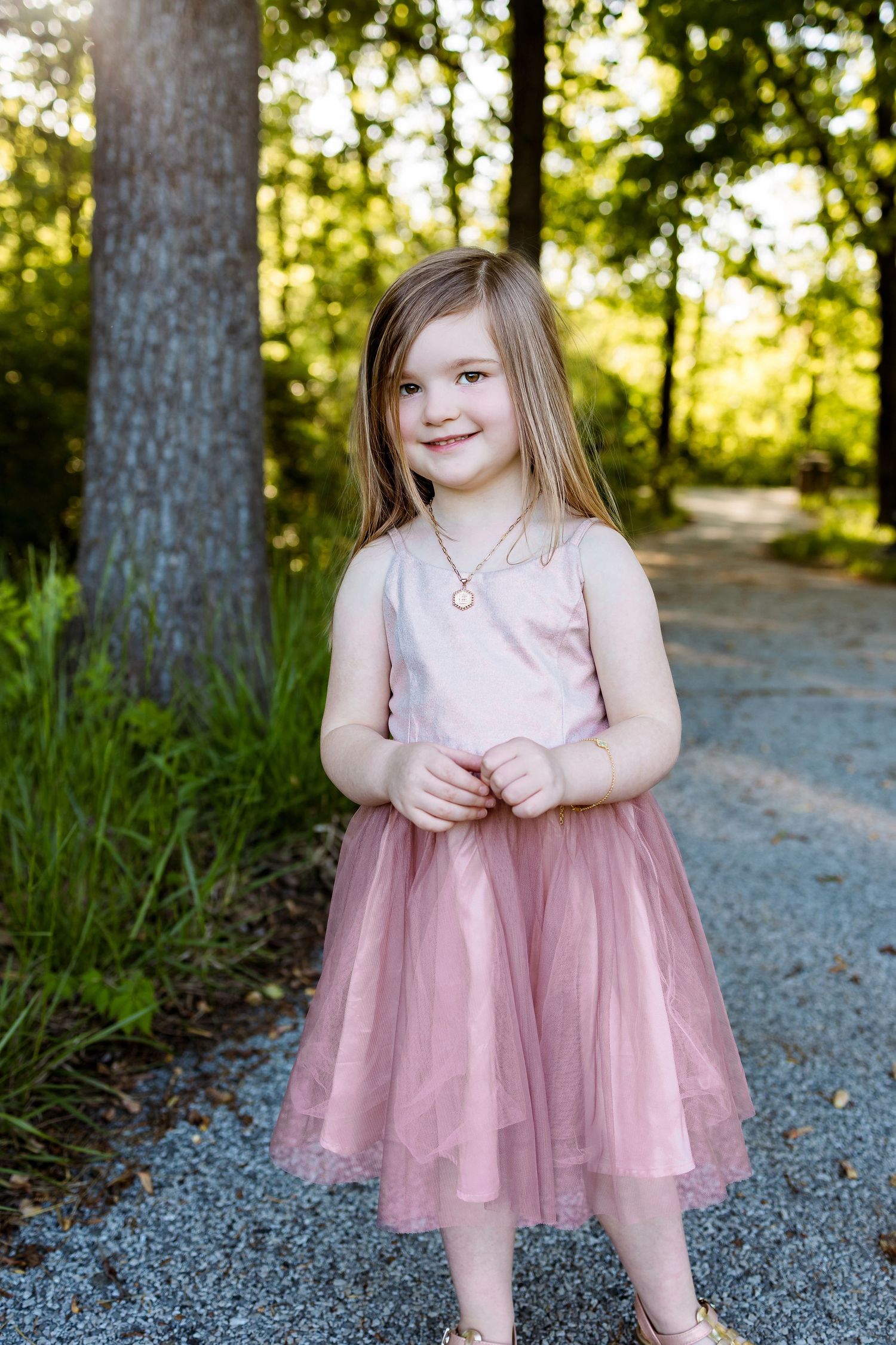 Young girl in a pink tulle dress poses sweetly on a wooded path during golden hour.