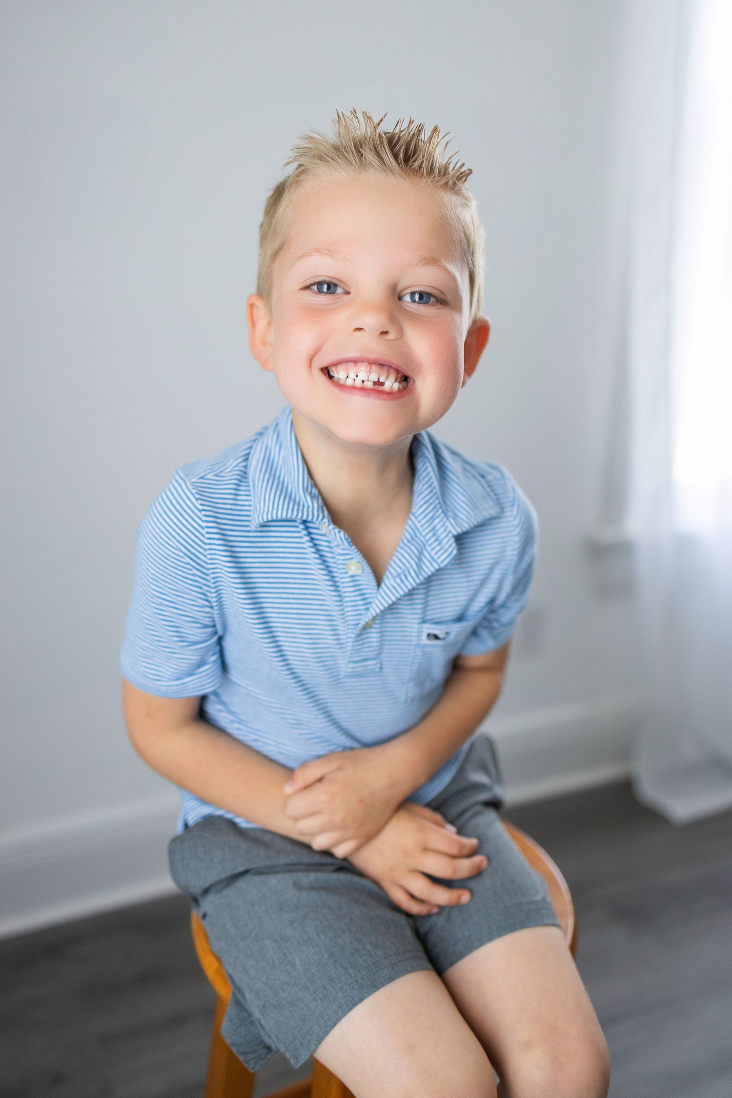 Child in blue striped polo shirt and gray shorts smiles while sitting on wooden stool.