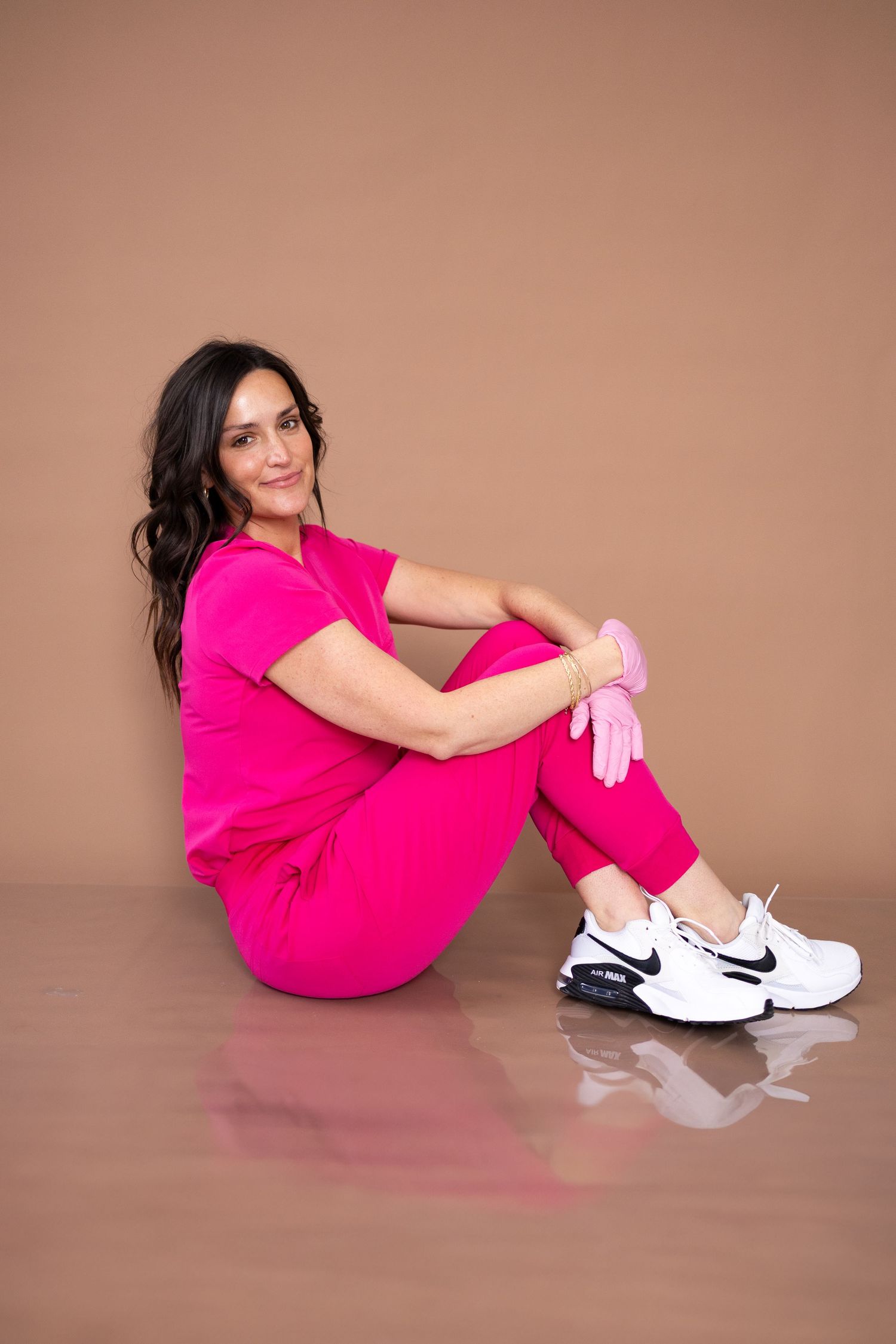 Person in pink medical scrubs and white sneakers sitting on reflective surface.