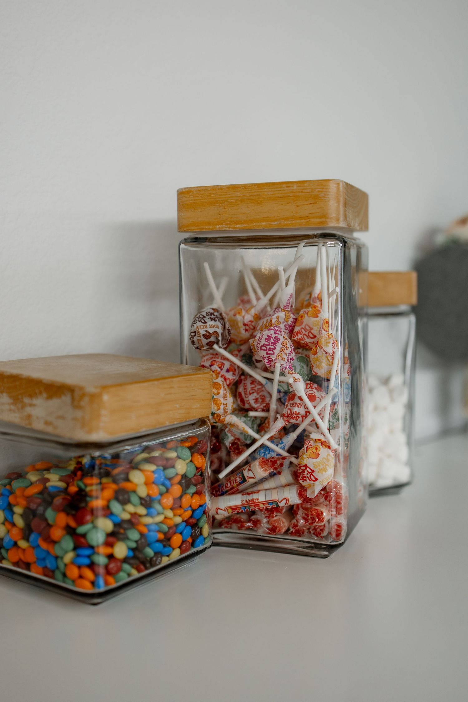 Glass jar with wooden lid containing colorful M&M candies and lollipops shown from multiple angles on white surface.