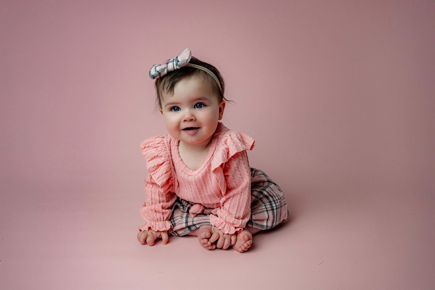 Baby wearing a peach-colored ruffled top and plaid pants poses on pink backdrop during professional photo session.