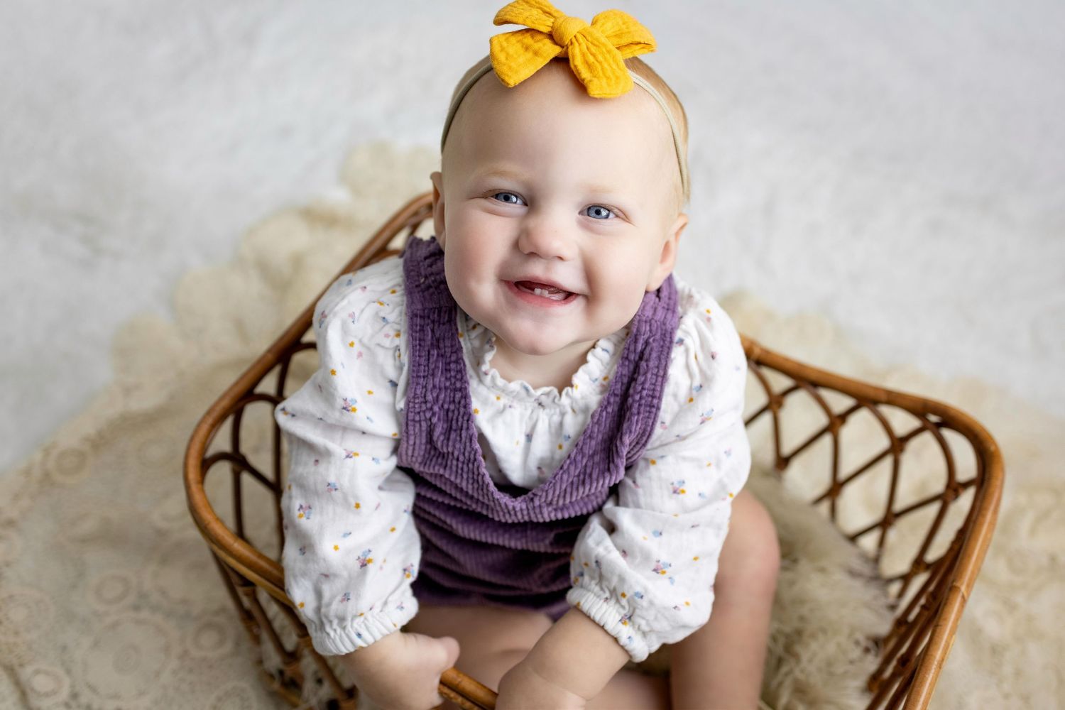 A small child in a purple dress and white lace top sits in a woven wicker chair against a neutral background.