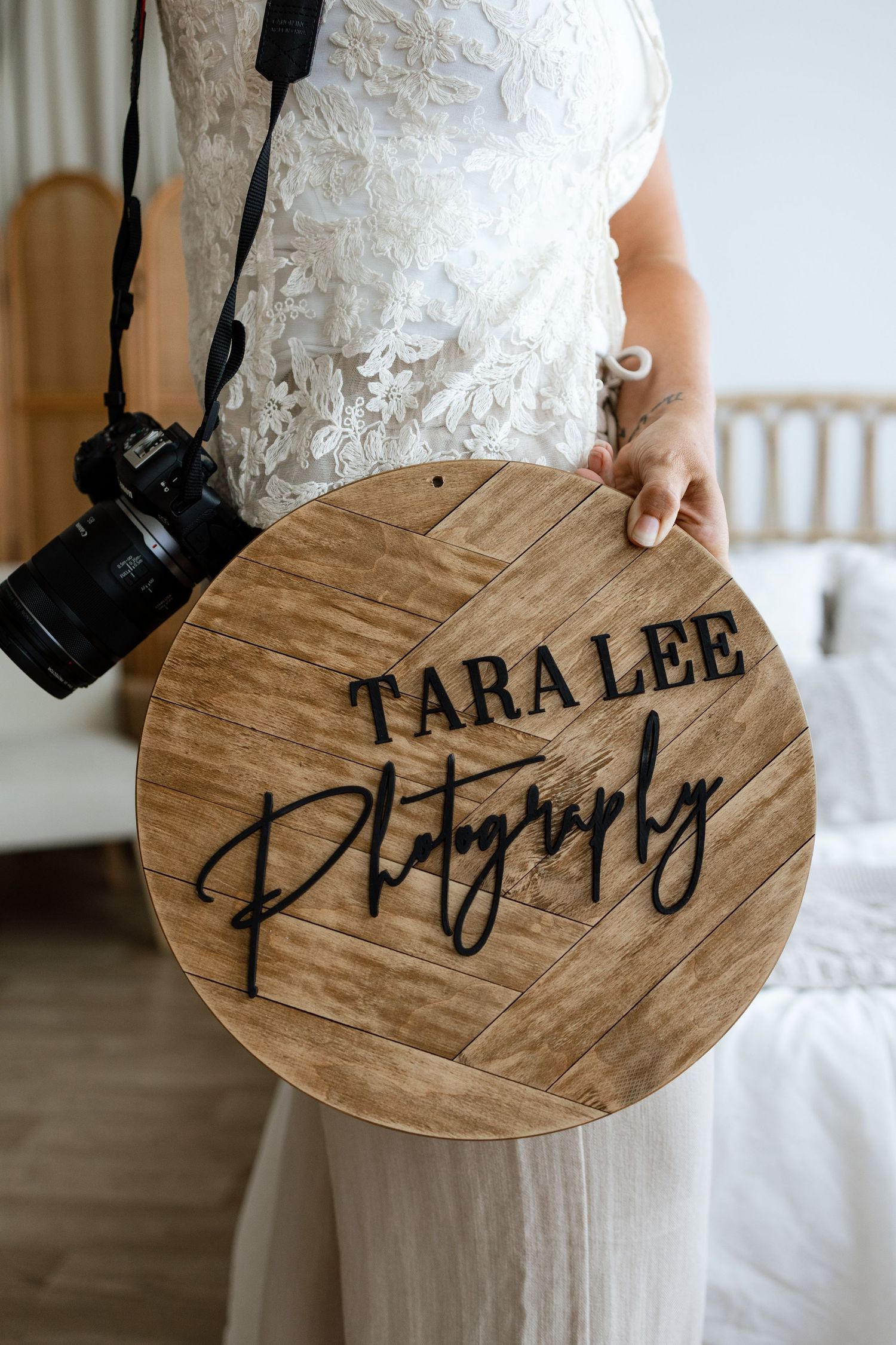 A bride in a white lace dress holds a round wooden Tara Lee Photography sign while wearing a camera strap.