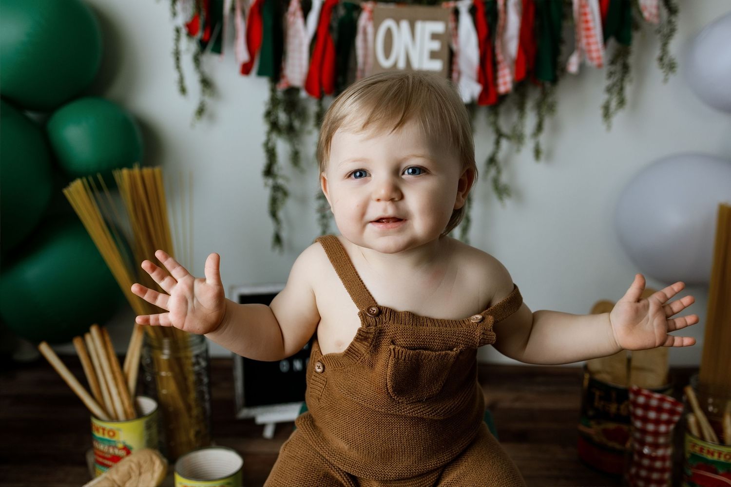 Toddler in brown overalls smiles at party with green balloons and festive banner decorations in background.