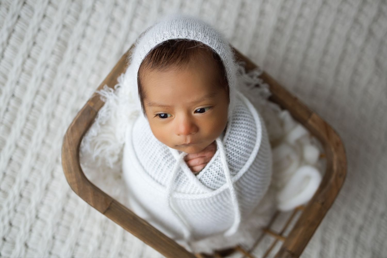 A baby wearing a white knit sweater lies on a textured cream-colored blanket.