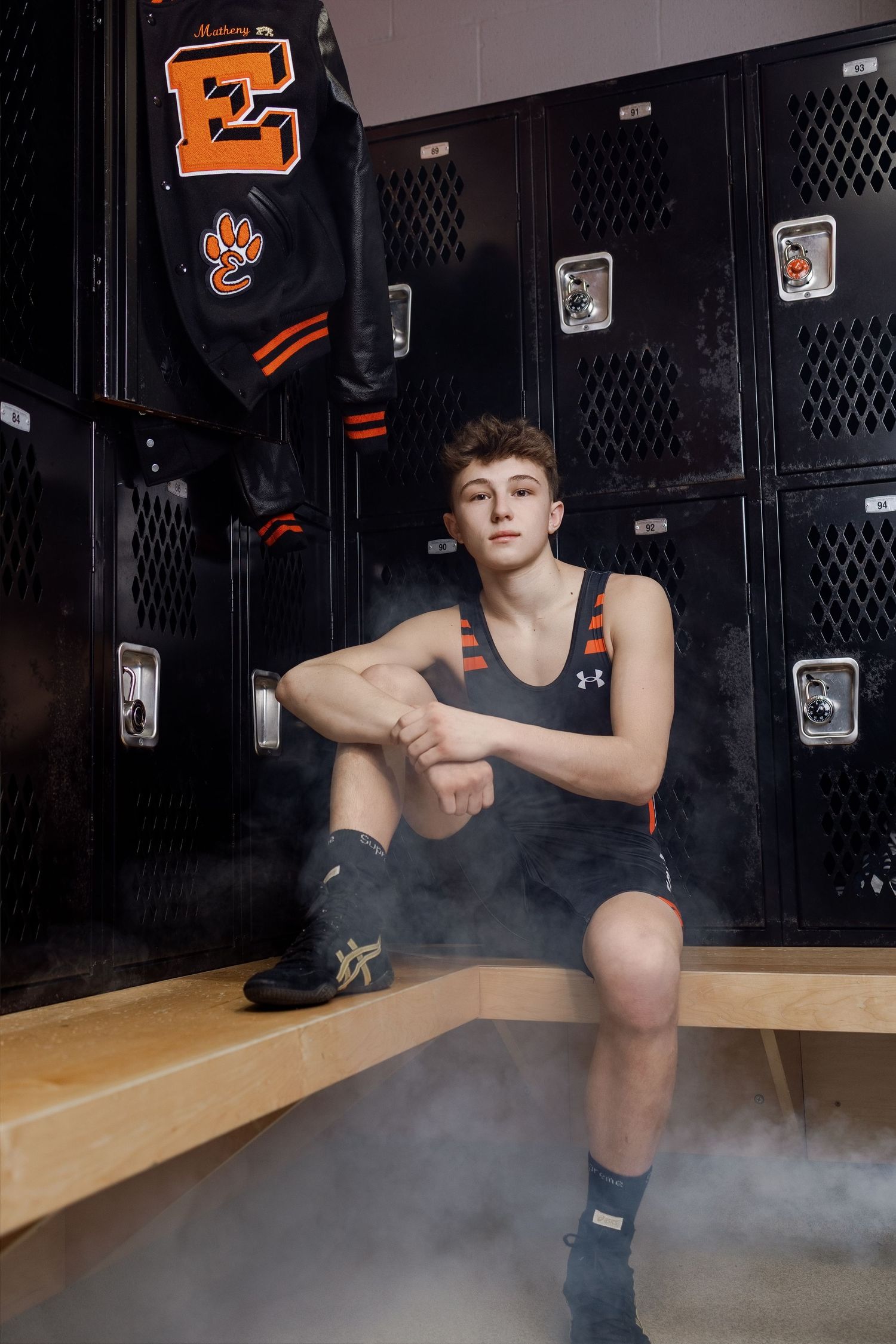 High school athlete in Under Armour gear sits on bench in front of lockers with orange varsity letter jacket visible.