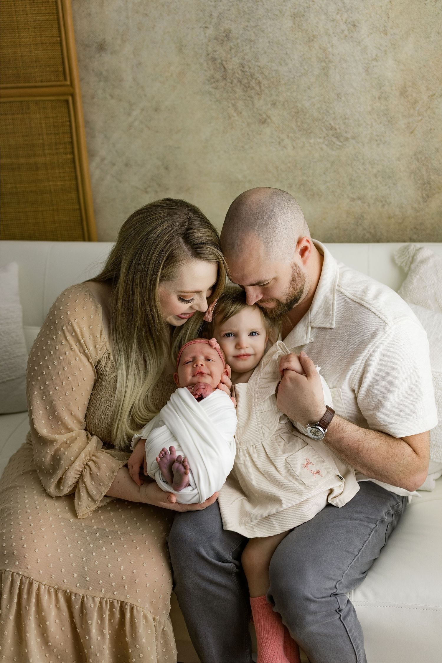 Intimate family portrait showing tender moment of parents cuddling together on light colored couch.