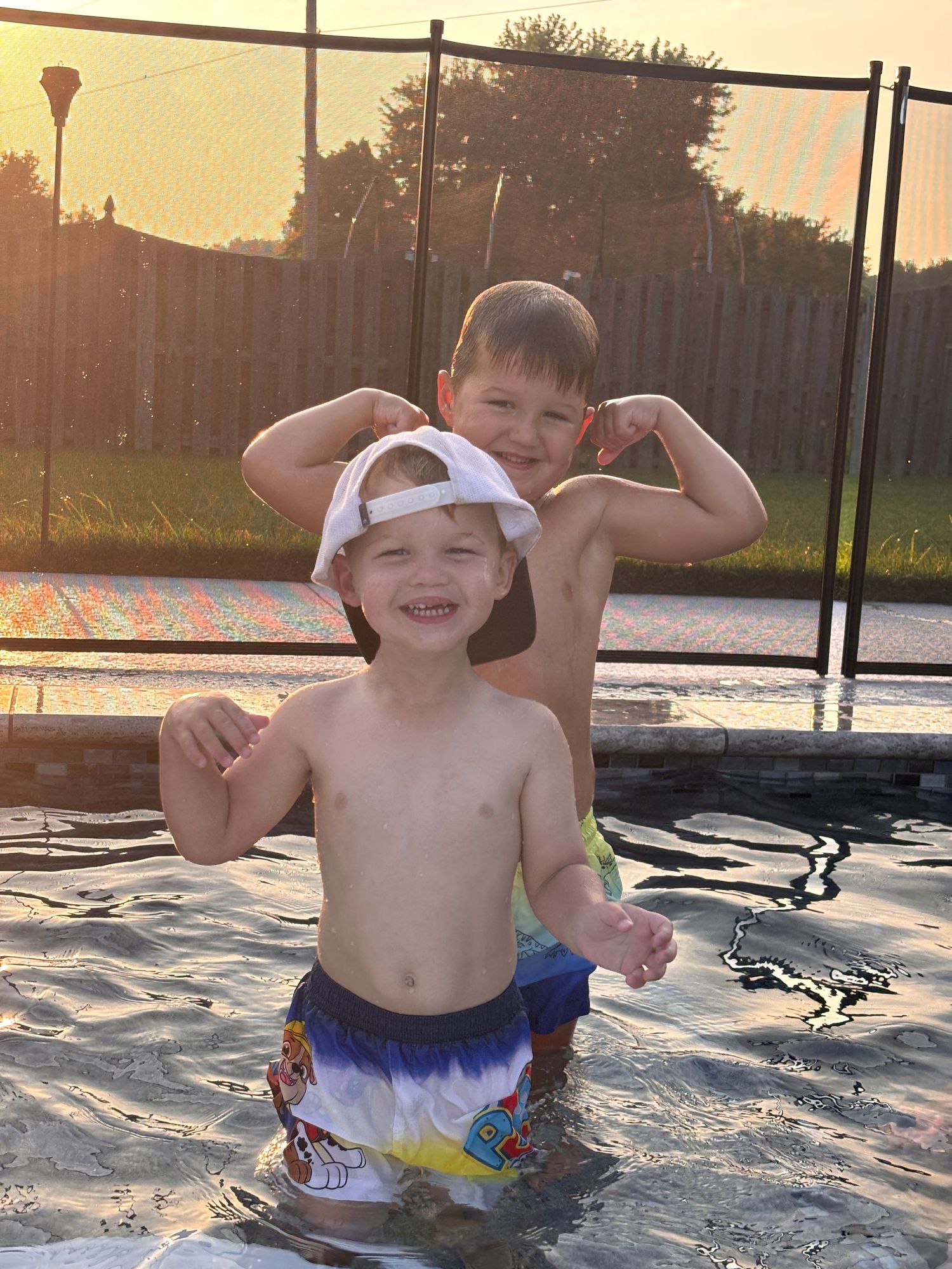 Two young boys flexing muscles in a backyard pool at sunset, wearing swim trunks and a white cap.