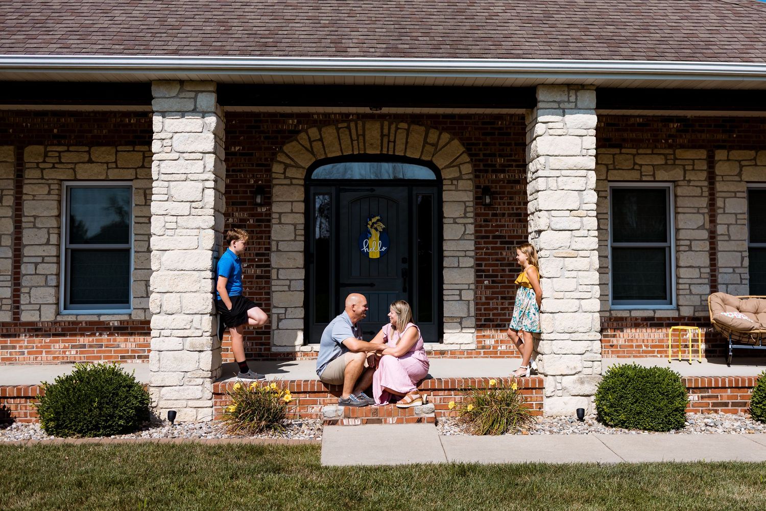 A stone facade ranch house with a covered front porch and landscaped bushes in sunlight.