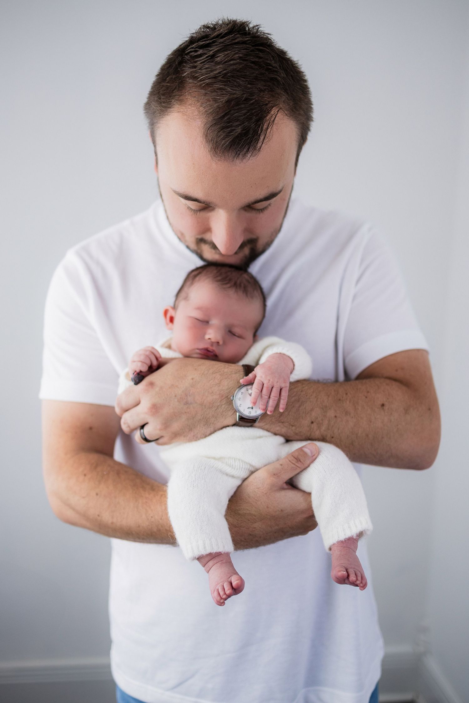 A tender moment between adult in white t-shirt and peaceful sleeping newborn baby against light gray background.