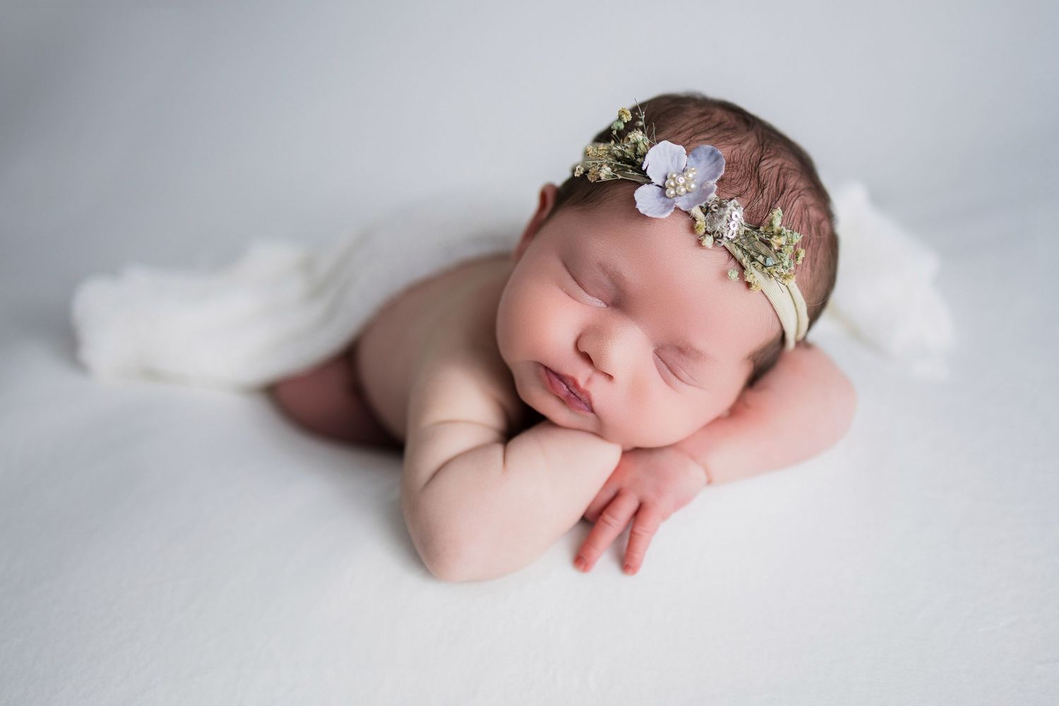 A sleeping newborn baby wearing a delicate floral headband rests peacefully on white fabric.