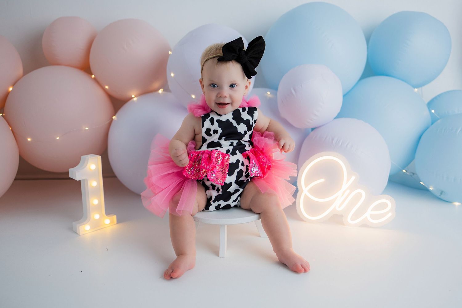 Baby feet with pink tutu in soft pastel nursery setting with blue and pink balloons and string lights in background.