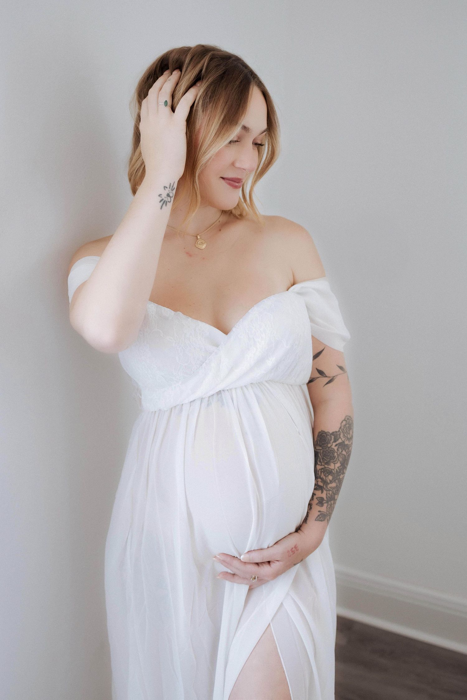 Expectant mother in white flowy dress poses against grey wall in elegant maternity portrait.