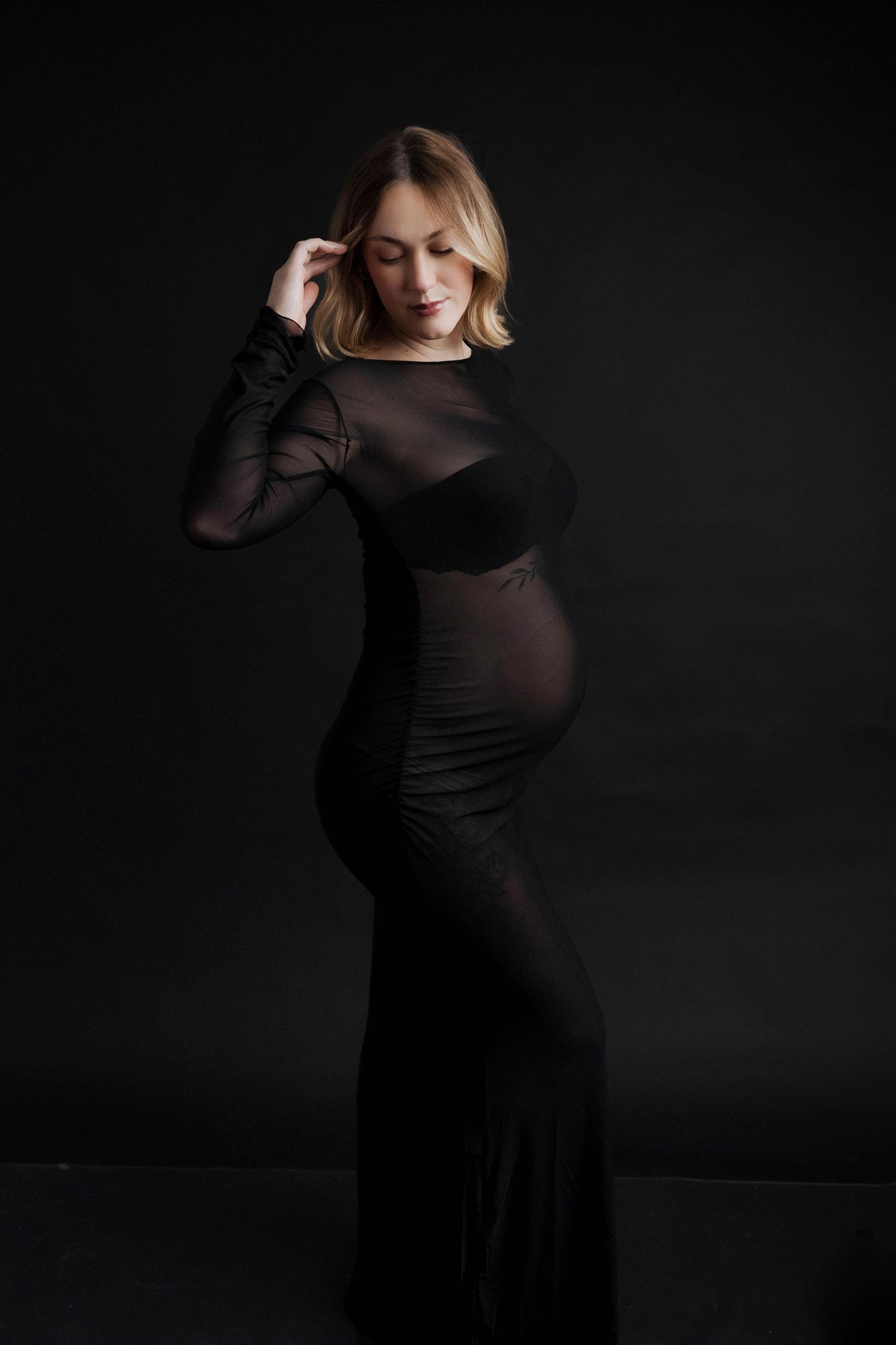 Elegant maternity portrait in black sheer dress against dark background showcasing pregnancy silhouette.