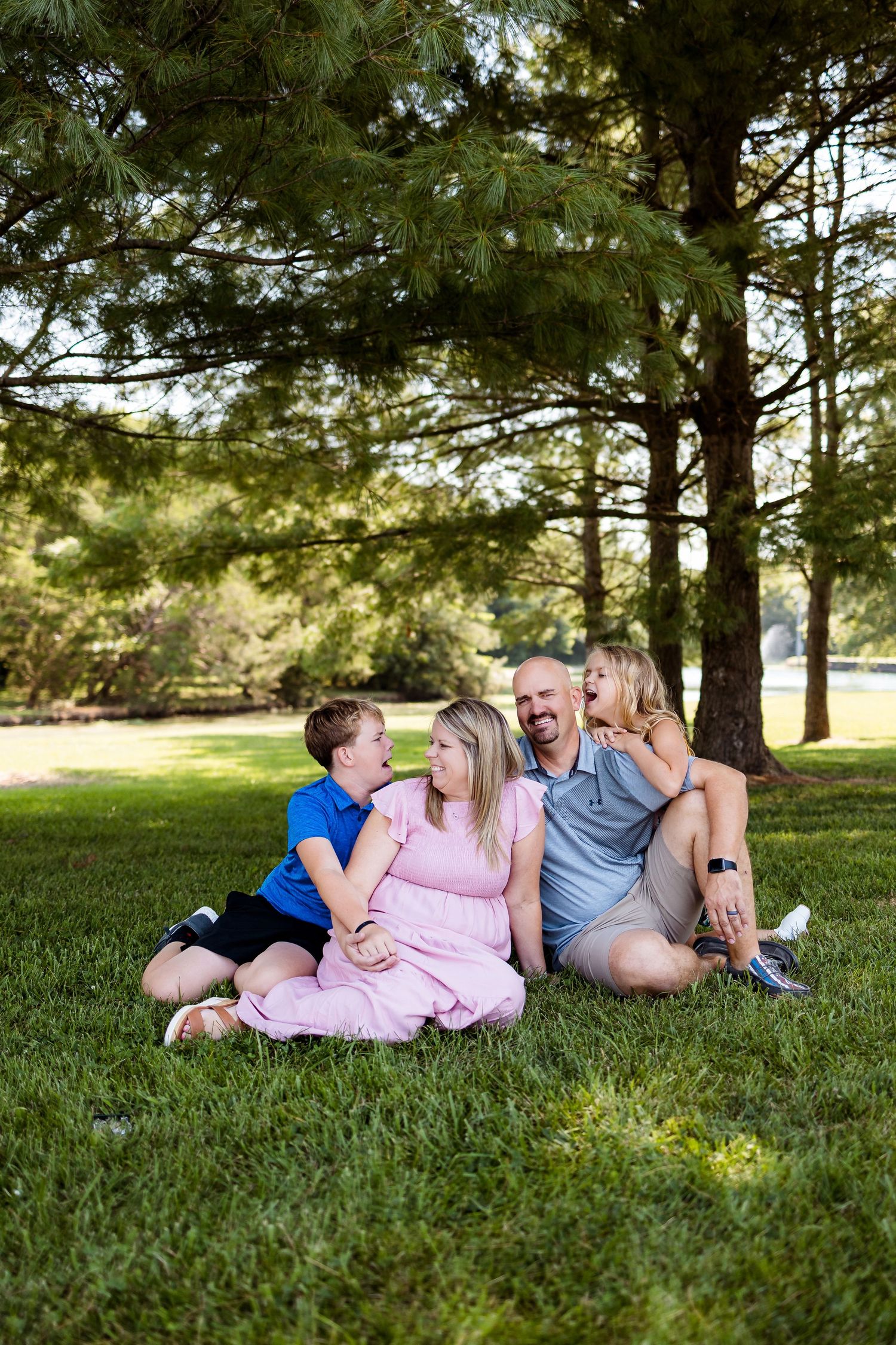 A family relaxes together on green grass under tall pine trees during a summer outdoor photo session.