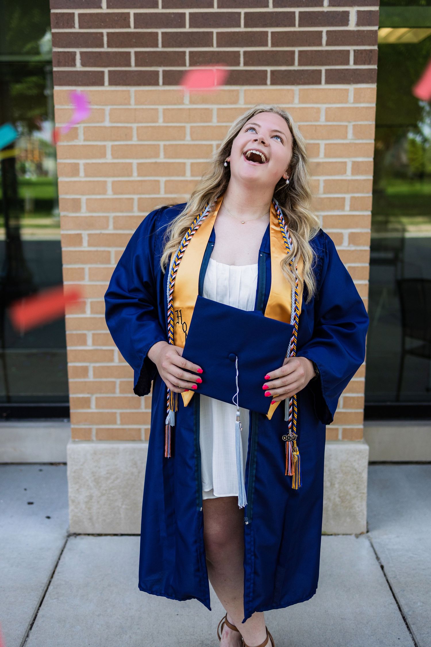 Graduate celebrates in blue graduation gown with gold honors stole while confetti falls against brick wall.