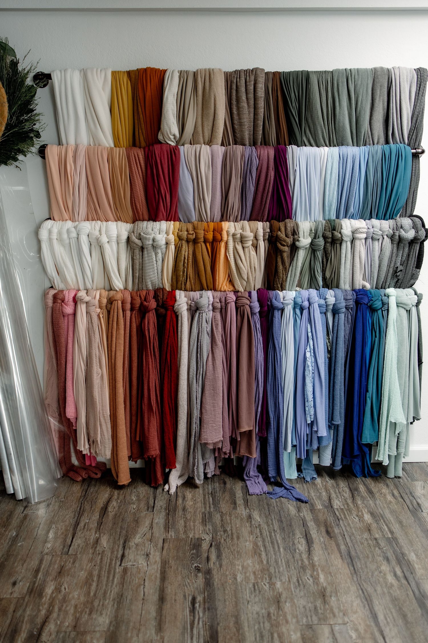 A collection of colorful bridesmaid dresses arranged by hue on hangers against a white wall with wooden flooring.