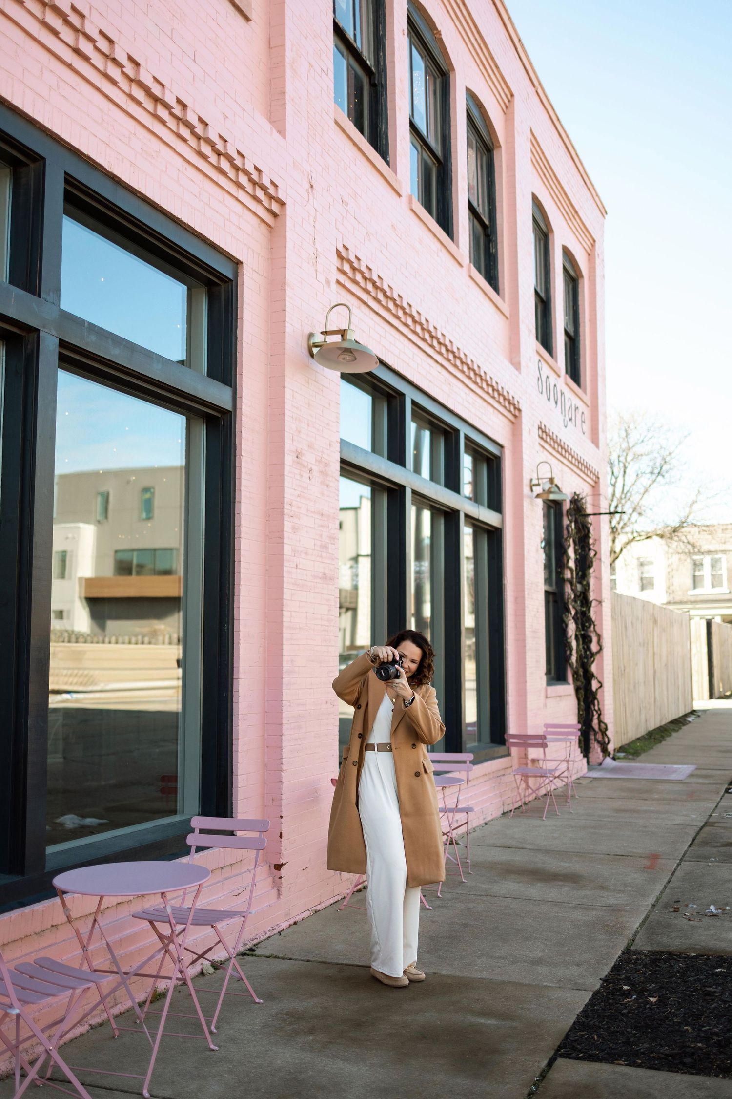 Person in white clothing poses along a pink building exterior with folding chairs and architectural details.