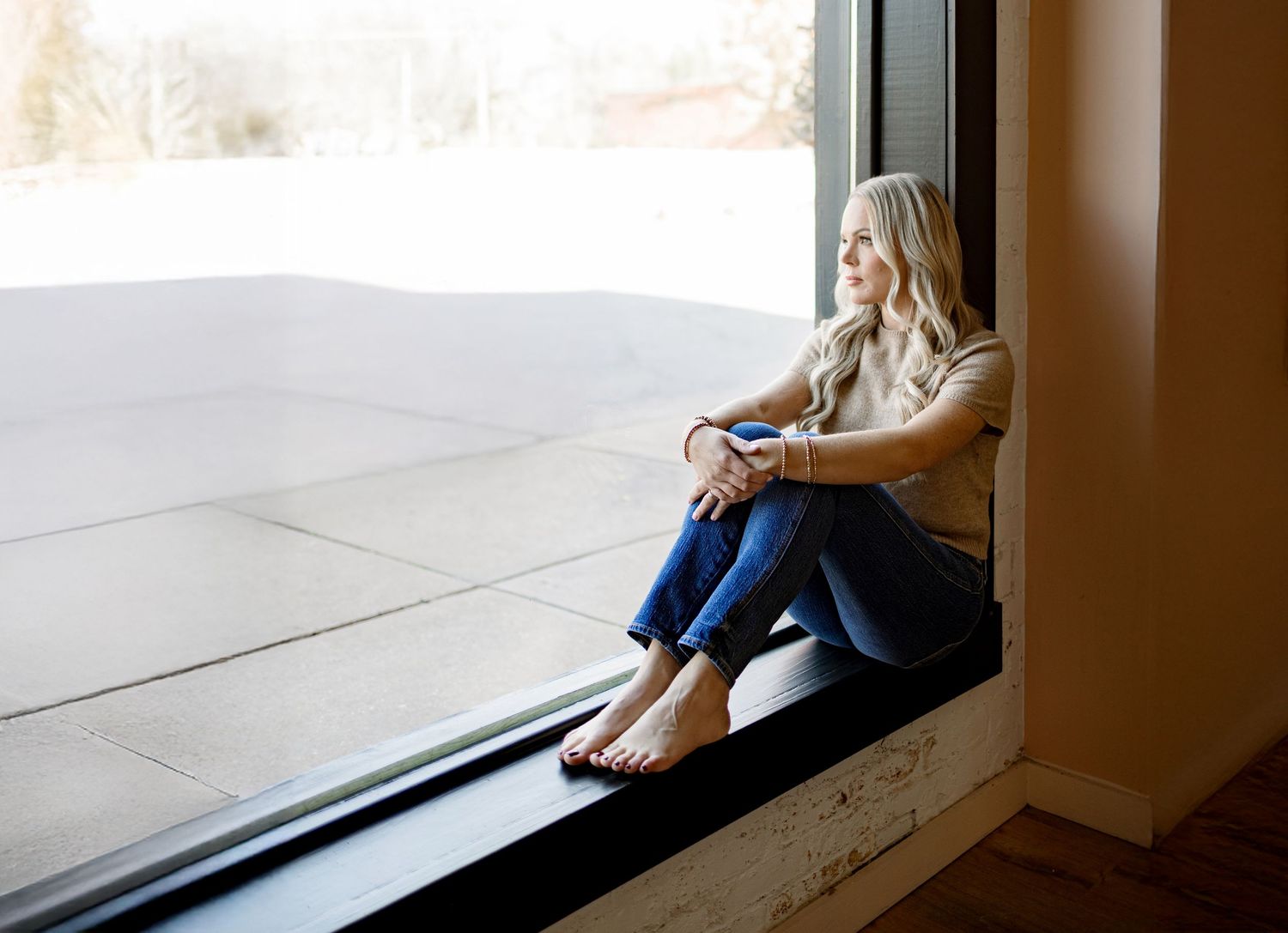 A woman in casual clothing sits alone by a large window, creating a contemplative mood in the minimalist space.