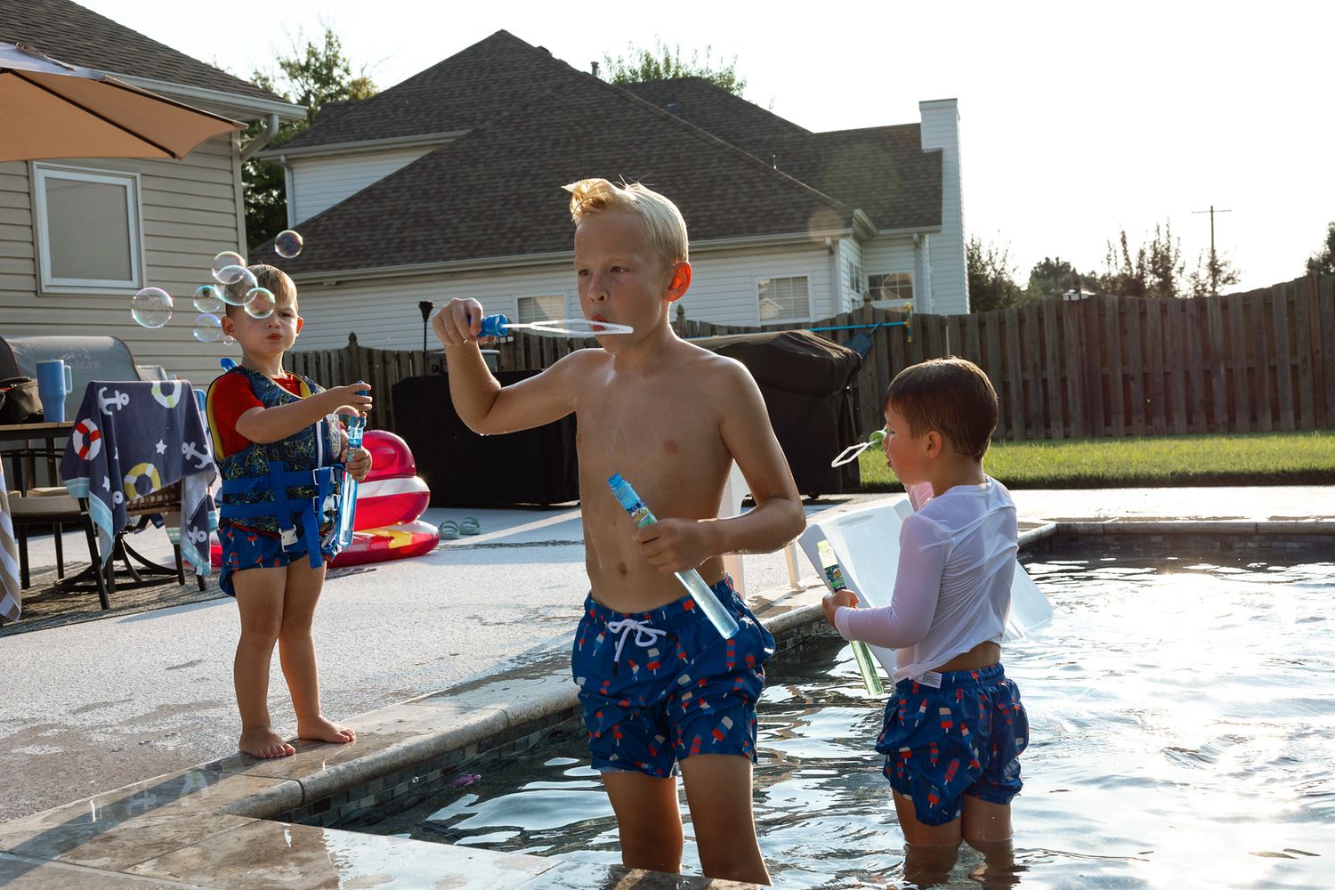 Three children playing with bubbles by a backyard swimming pool on a sunny summer day.