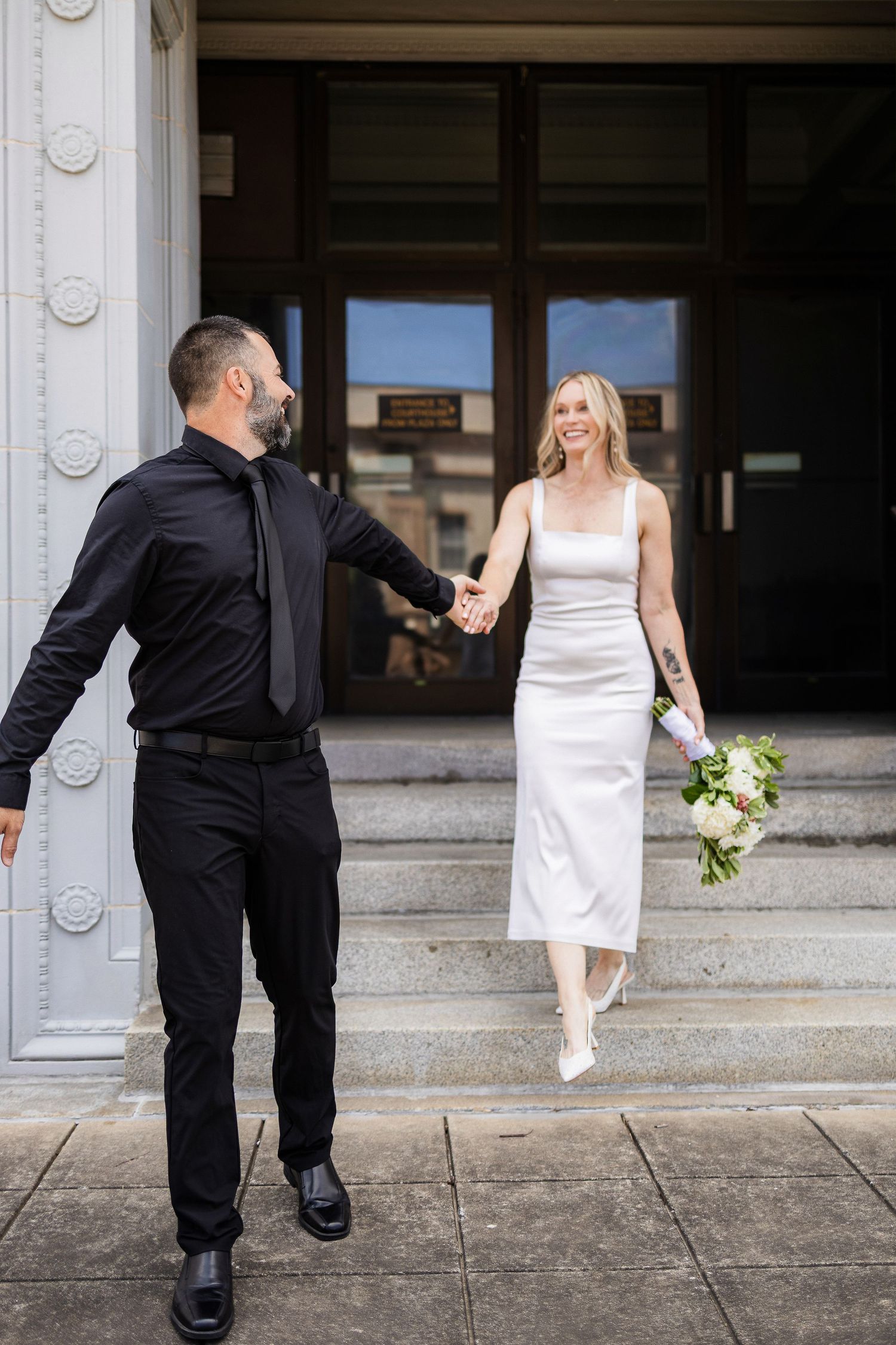 A couple in wedding attire descend stone steps together, with the bride in white holding a bouquet and groom in black.