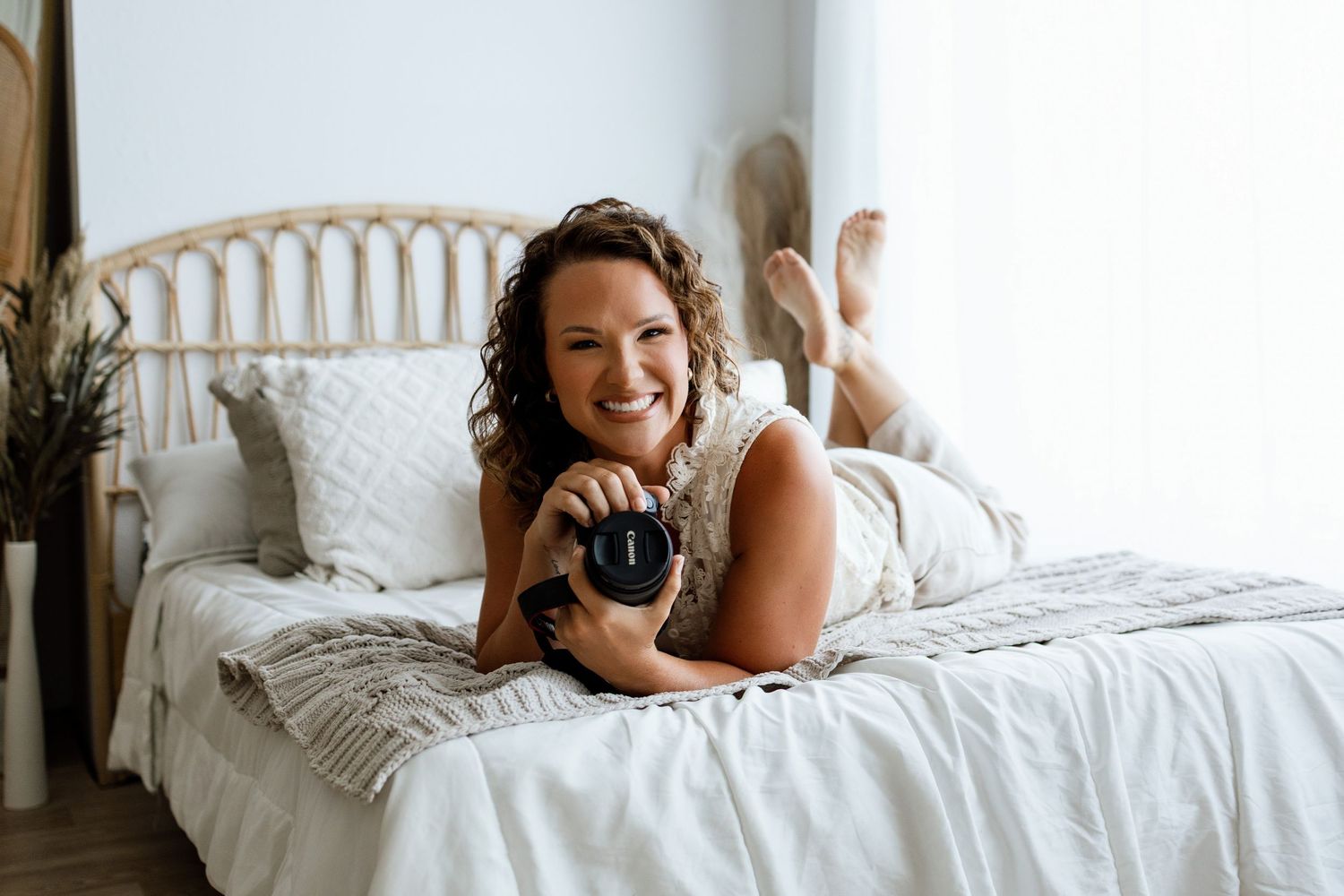 Woman with curly hair lying on white bed holding camera, smiling in bright boho bedroom.