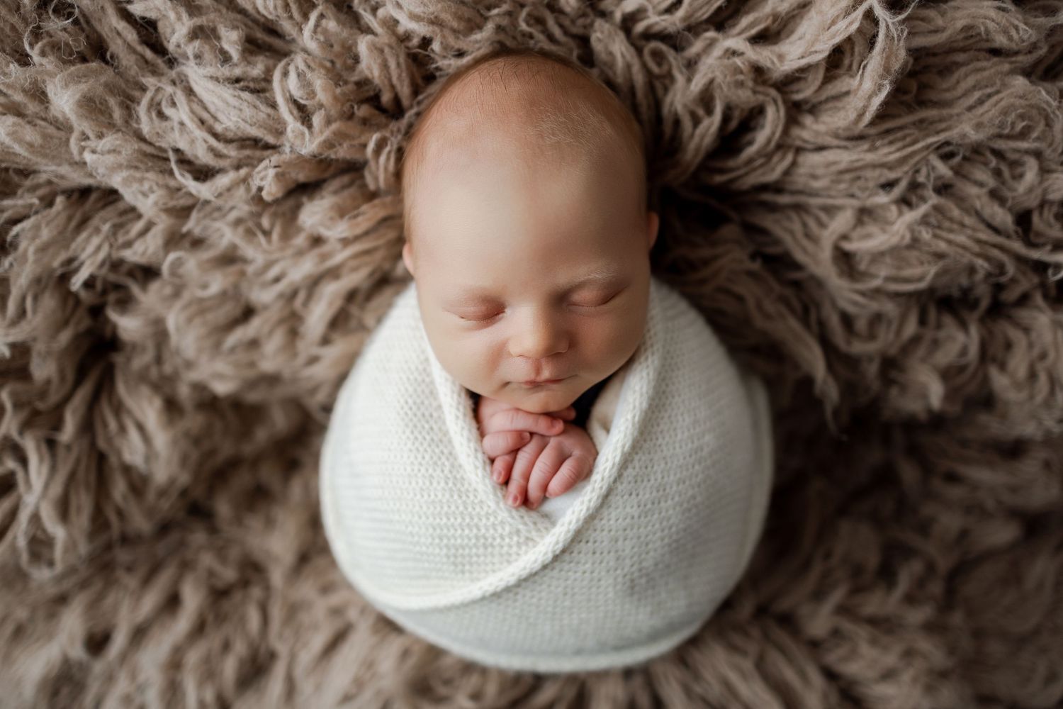 Sleeping newborn swaddled in white wrap on textured beige fur blanket photographed from above.