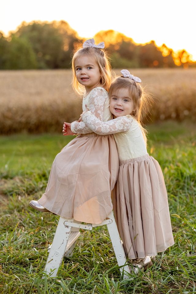 Two young children in matching lace dresses embrace while standing in a golden field at sunset.