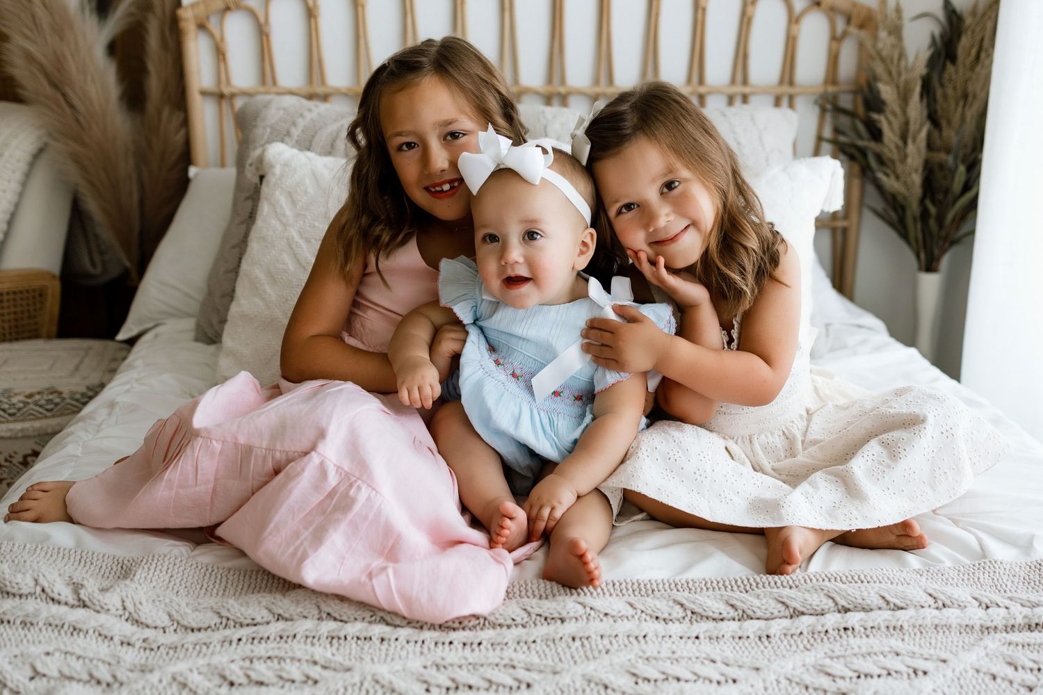 Two older sisters cuddle a smiling baby in blue on a cozy white bed, all dressed in pastel outfits.
