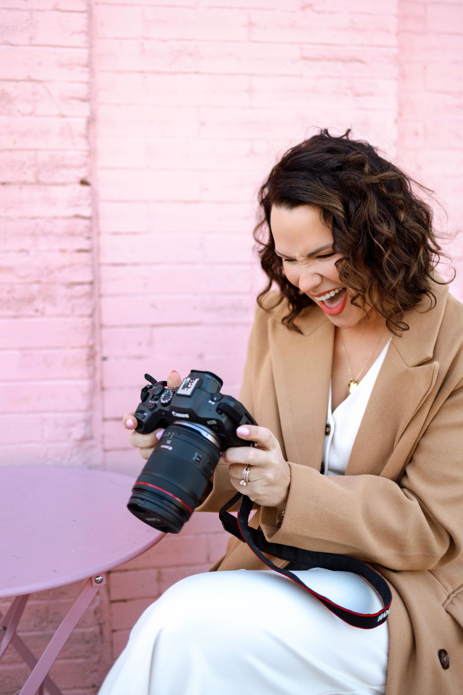 Photographer in beige coat laughs while reviewing photos on digital camera against pink wall.
