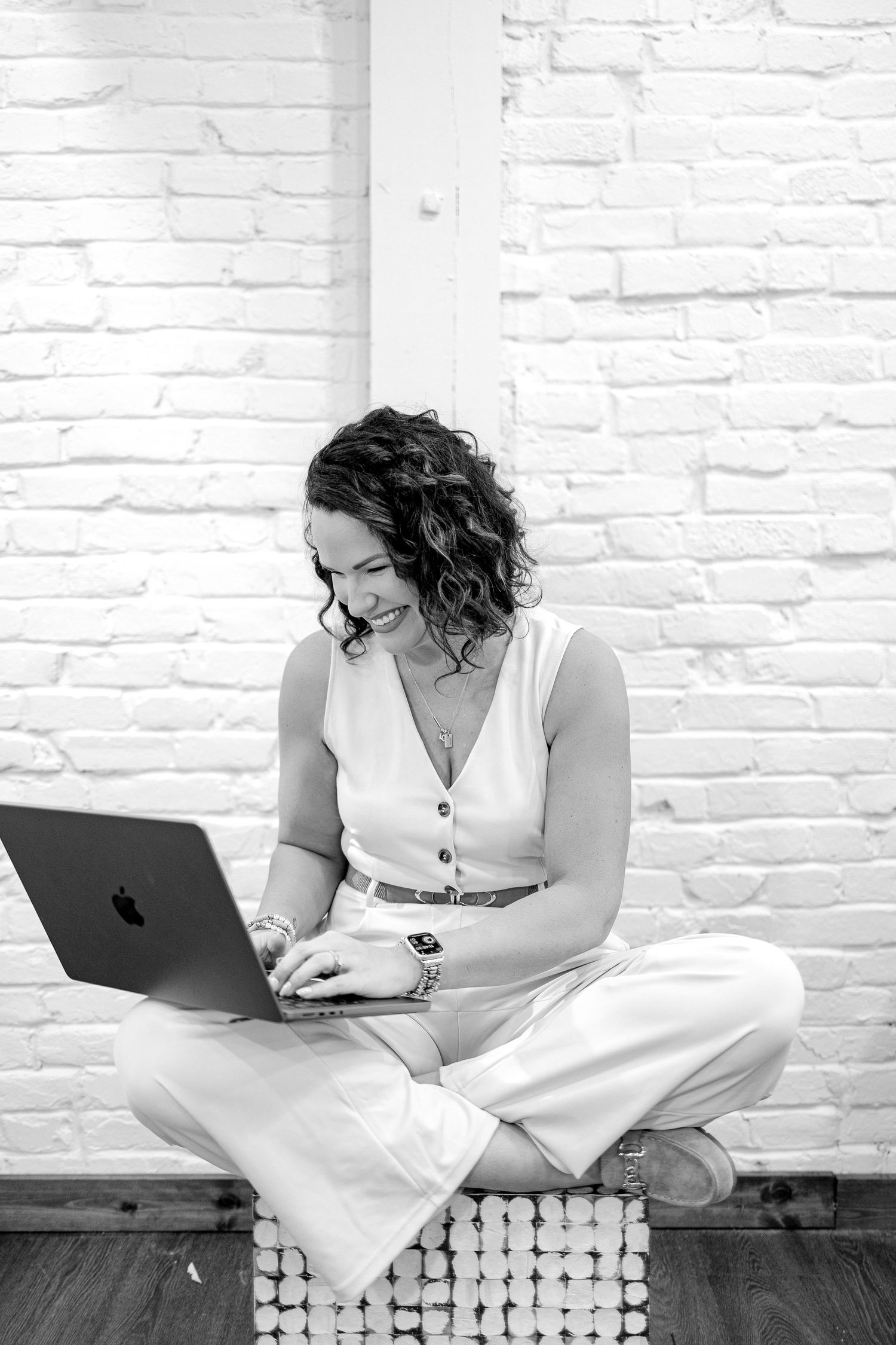 A person in a white dress sits on a wooden bench working on a laptop against a white brick wall.