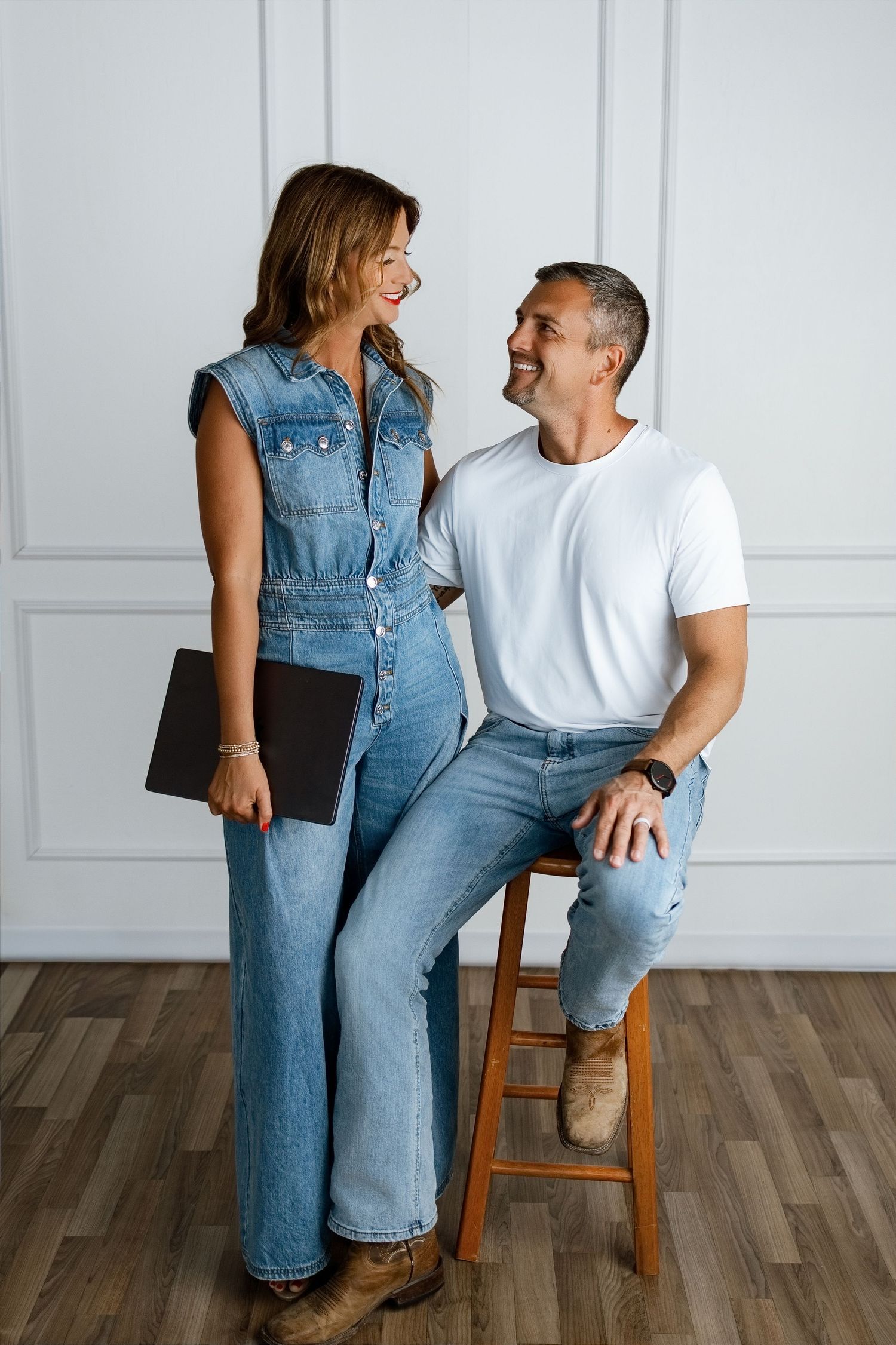 A smiling couple in casual denim outfits pose together in a bright white studio, woman holding a notebook.