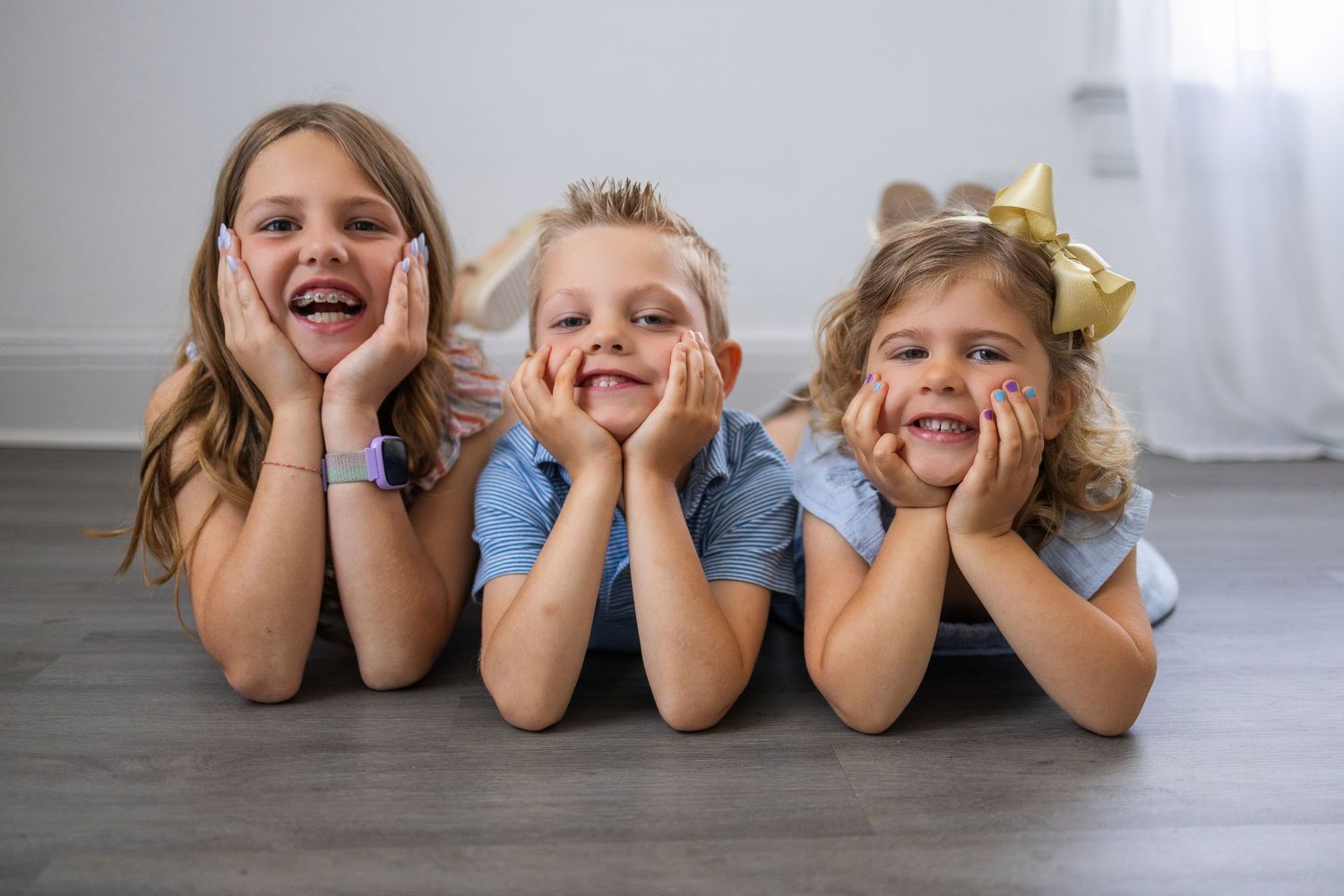 Three children lying on wooden floor with hands on cheeks smiling joyfully at camera.