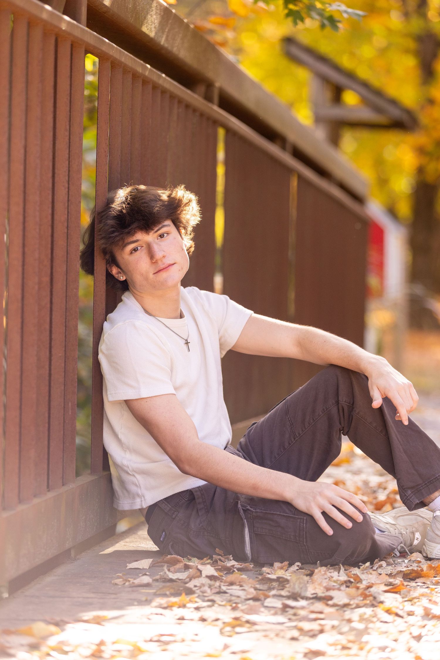 Casual autumn portrait with person sitting against wooden fence surrounded by fallen leaves.
